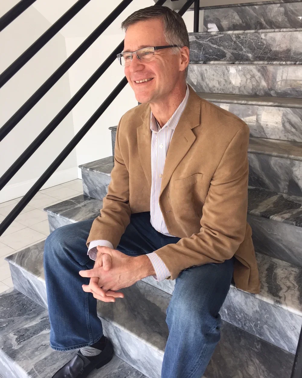 A smiling man with glasses, wearing a tan blazer and striped shirt, sitting on marble stairs with black railing in a modern indoor setting.