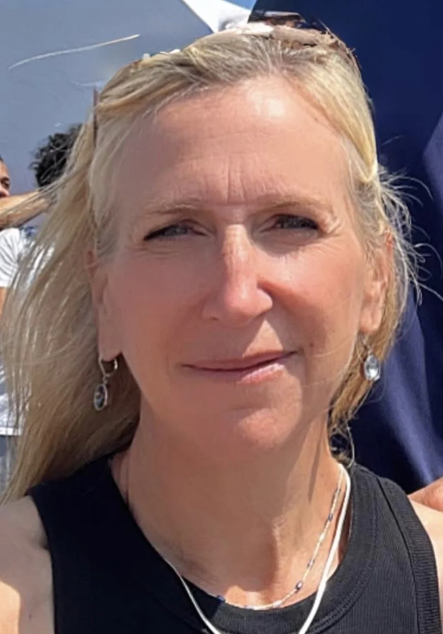 Close-up of a woman with blonde hair, wearing earrings and a black top, outdoors on a sunny day.