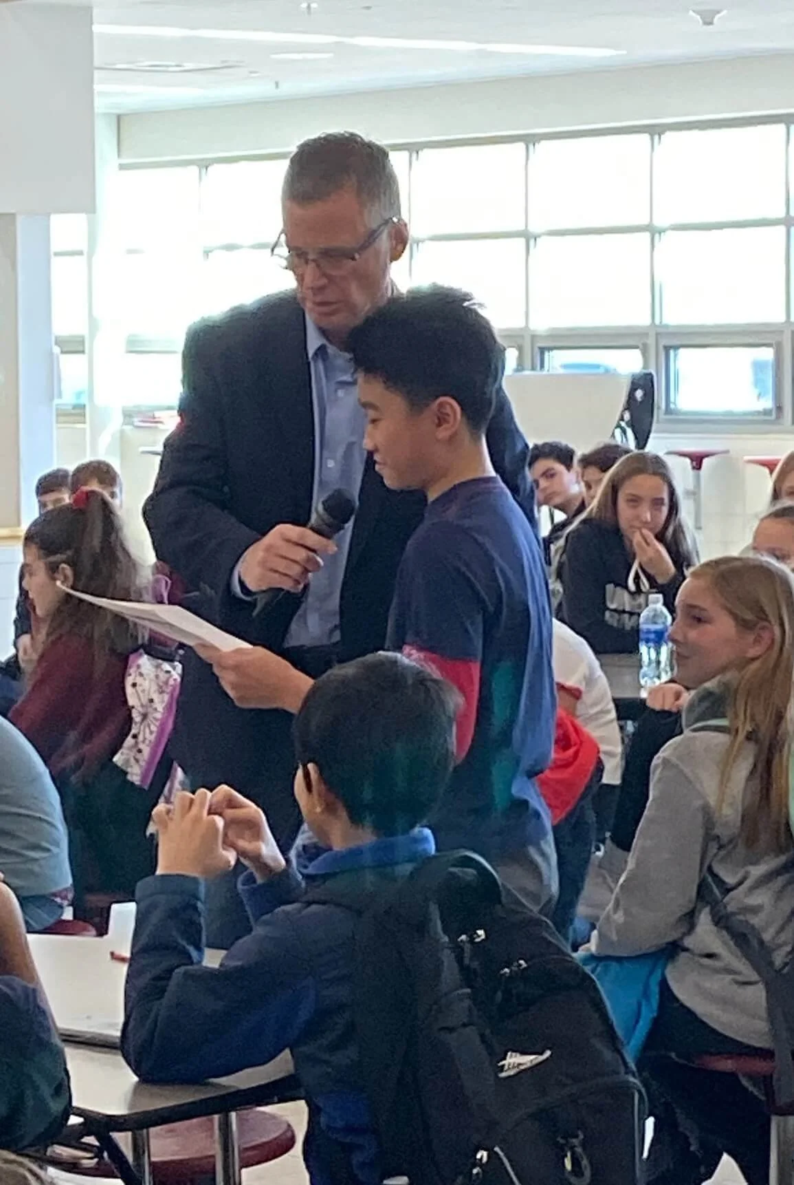 A man in a suit and glasses is holding a microphone and reading from a paper to a young student who is standing beside him at a school assembly. The assembly room is filled with students seated at tables, some are smiling and listening attentively.