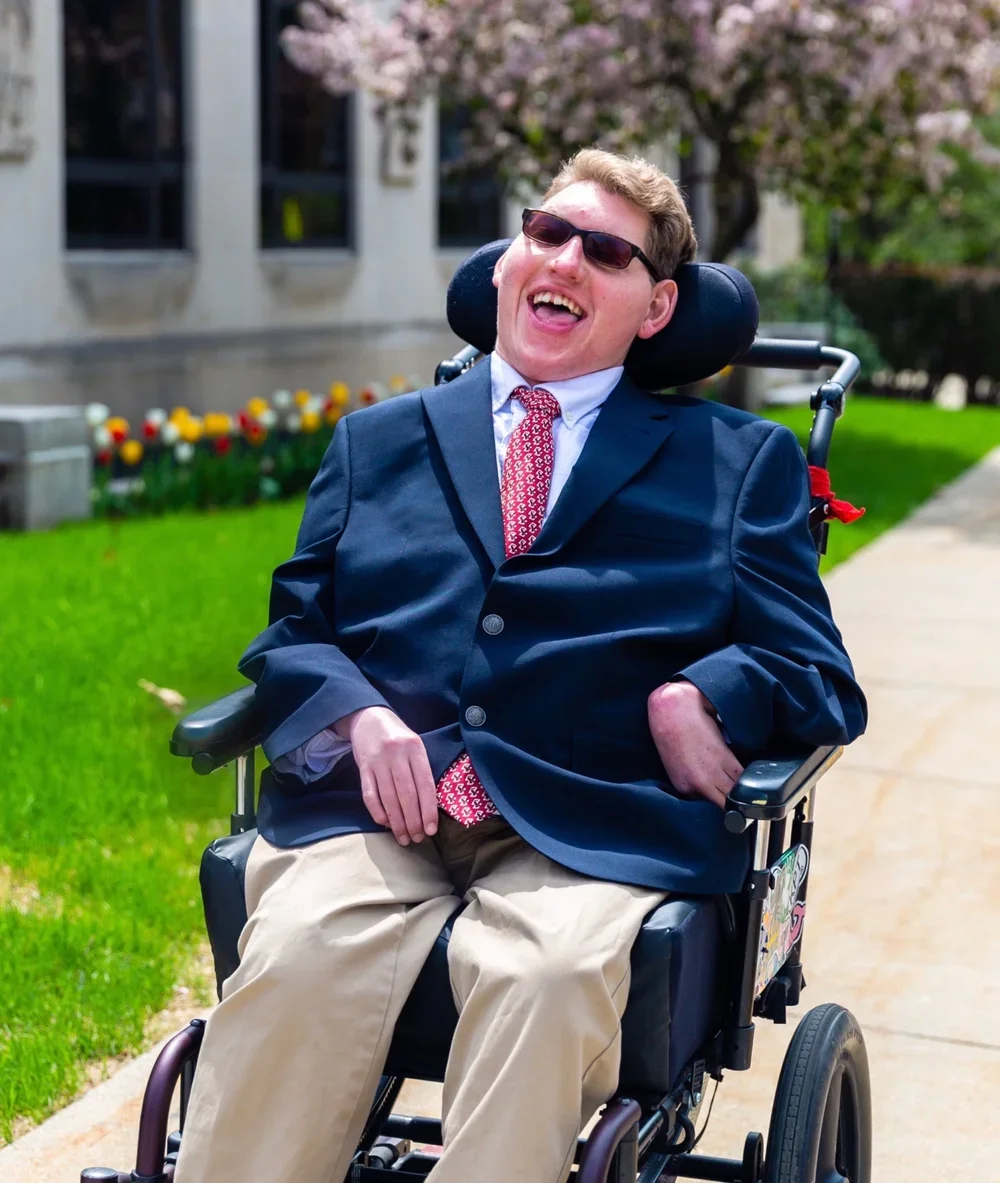 A smiling man in a wheelchair wearing sunglasses, a navy blazer, a white shirt, a red patterned tie, and tan pants, sitting outdoors on a sidewalk with a building and pink flowering trees in the background.