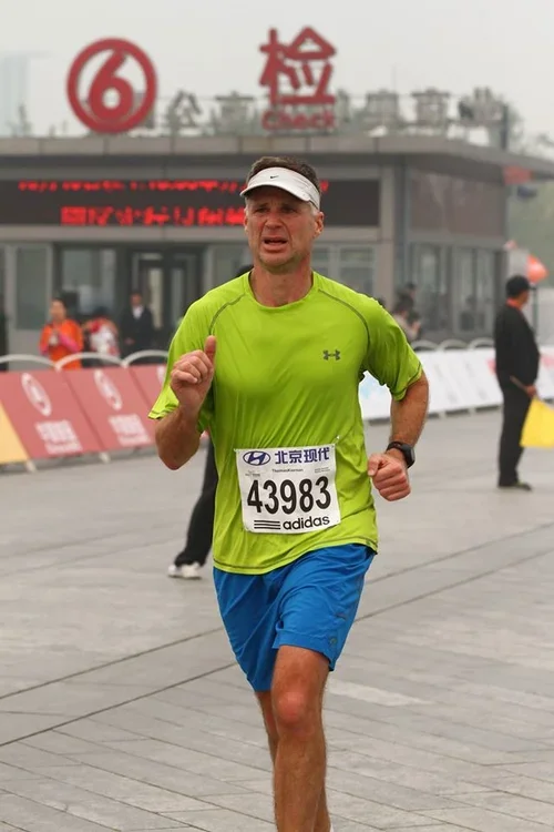 A male runner in a bright green shirt and blue shorts participating in a race, with a race bib numbered 43983, running outside a building with Chinese signage and a red number 6 logo.
