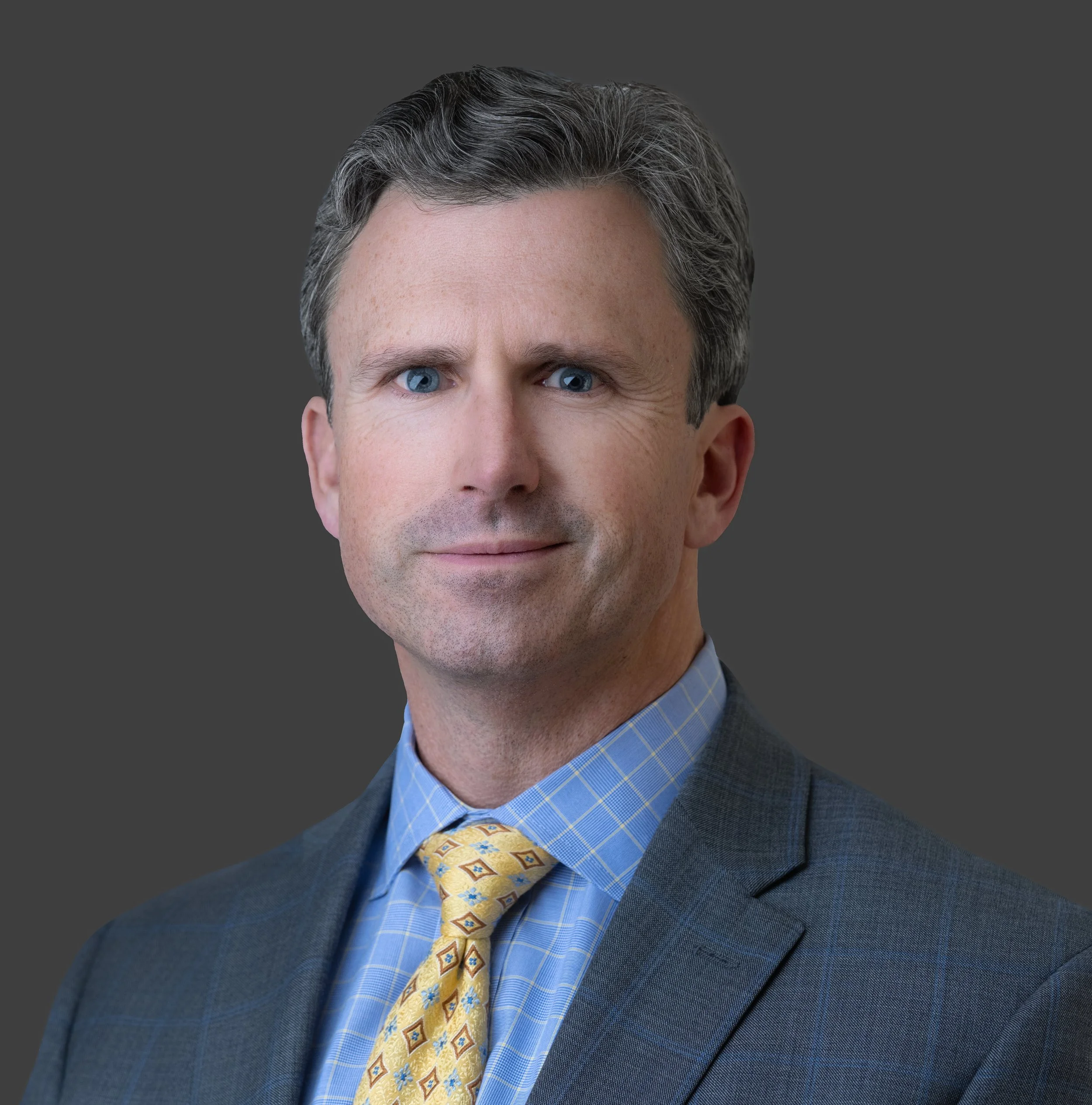 Close-up of a middle-aged man's face with blue eyes, gray hair, and wearing a dark blue suit, light blue checkered shirt, and yellow patterned tie, against a dark gray background.