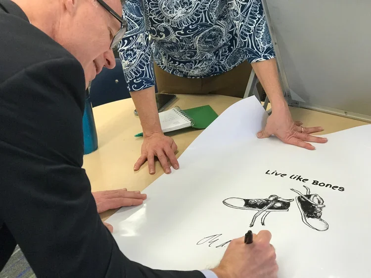 Two people with one signing a poster that has a drawing of a cockroach and the text "Live like Bones," on a desk.