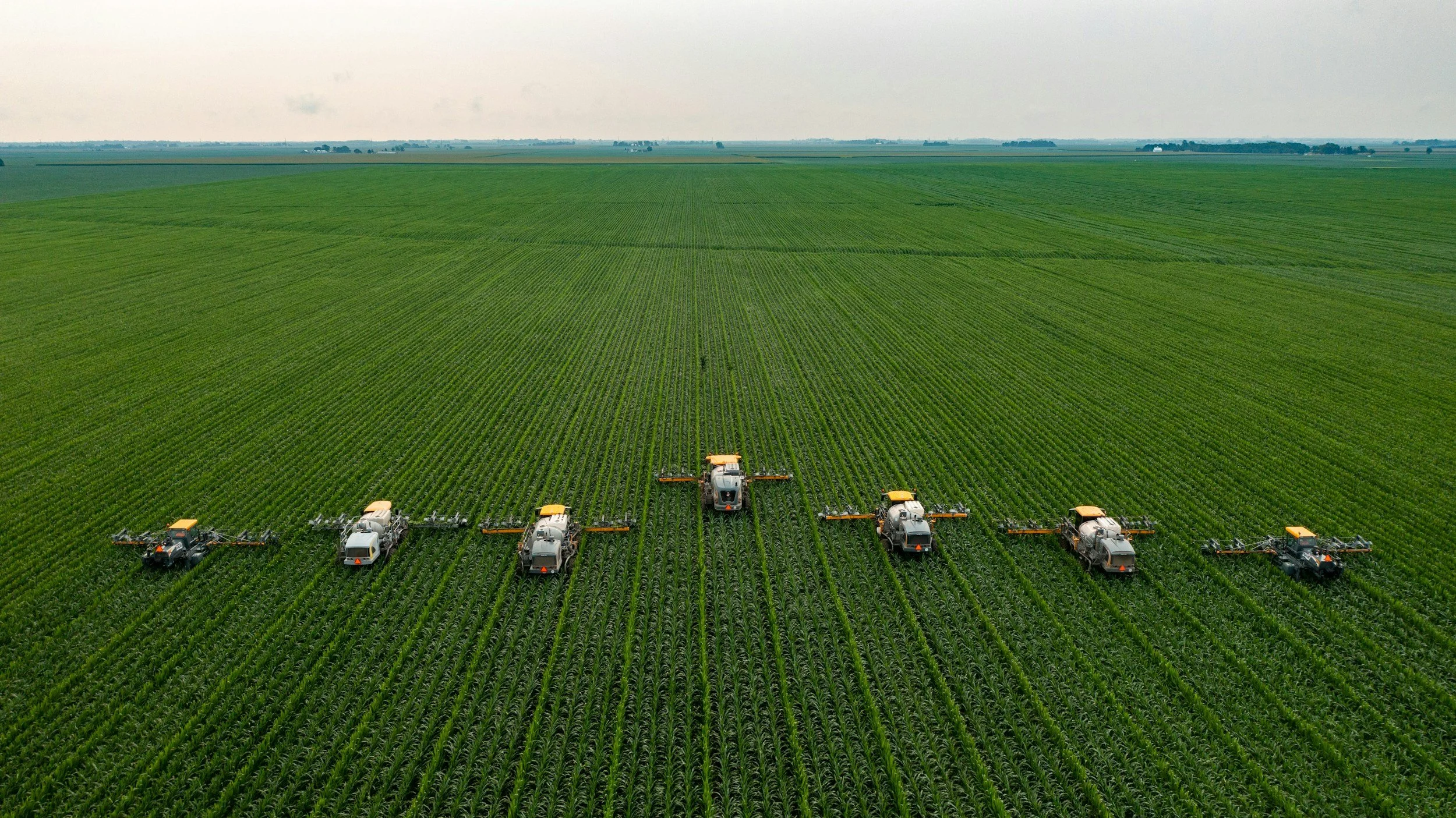Six agricultural spraying machines working on a large green crop field from above.