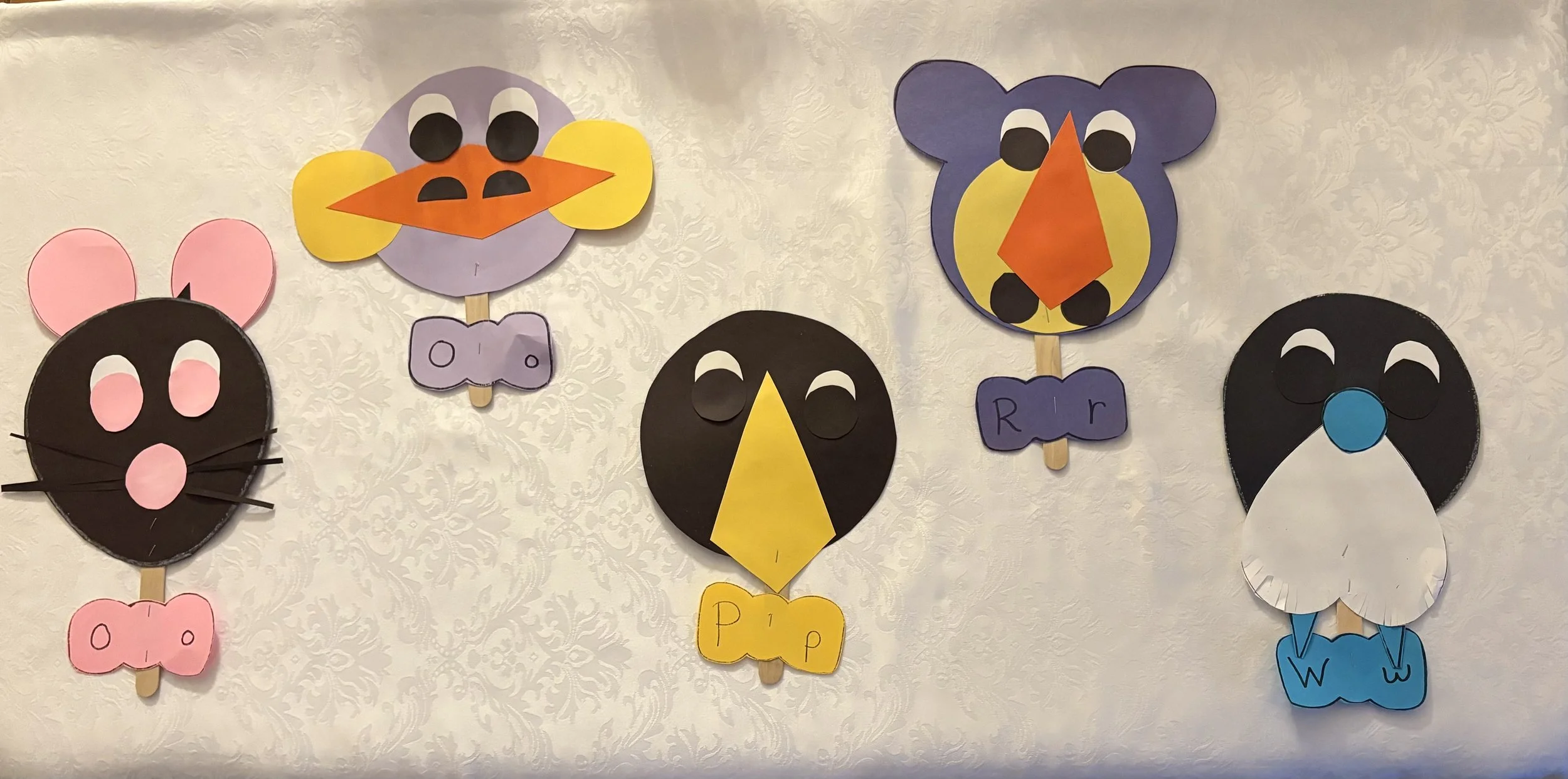 Colorful paper puppets of various animals, including a pig, a pig with pink ears, a bird, an owl, and a penguin, attached to sticks and displayed on a white wall.