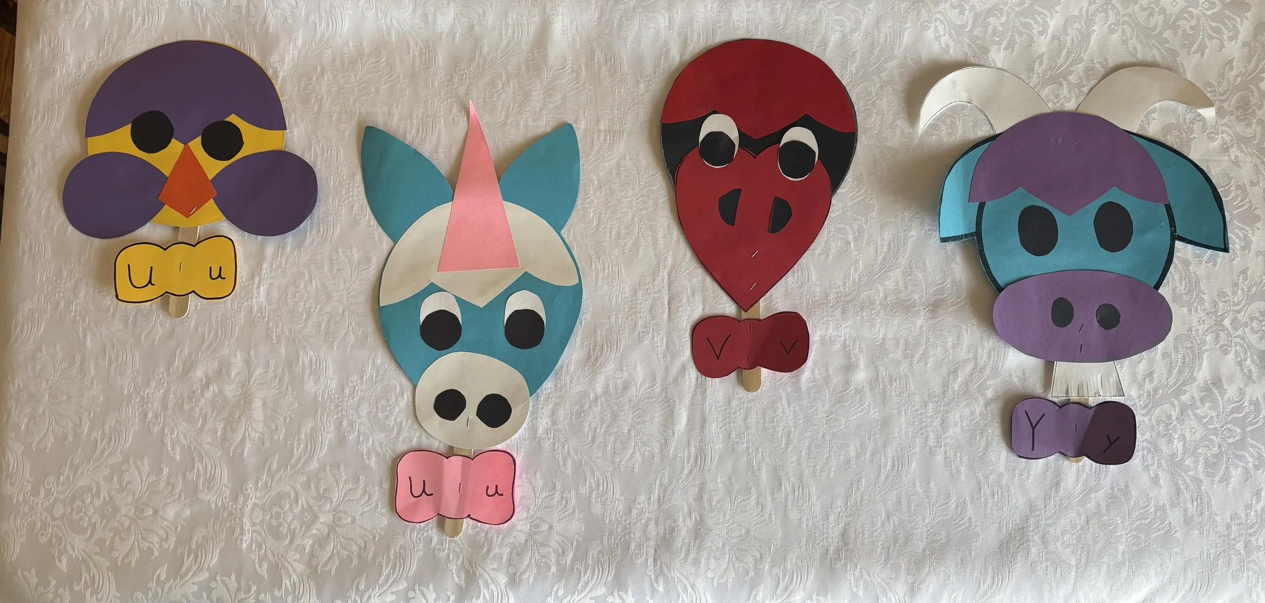 Colorful paper masks of animals and characters displayed on a white textured tablecloth. From left to right, a purple owl with yellow eyes and an orange beak, a blue and white rabbit with pink nose and ears, a red pig with black details, and a purple and blue dinosaur with silver horns. Each mask has a stick at the bottom for holding.