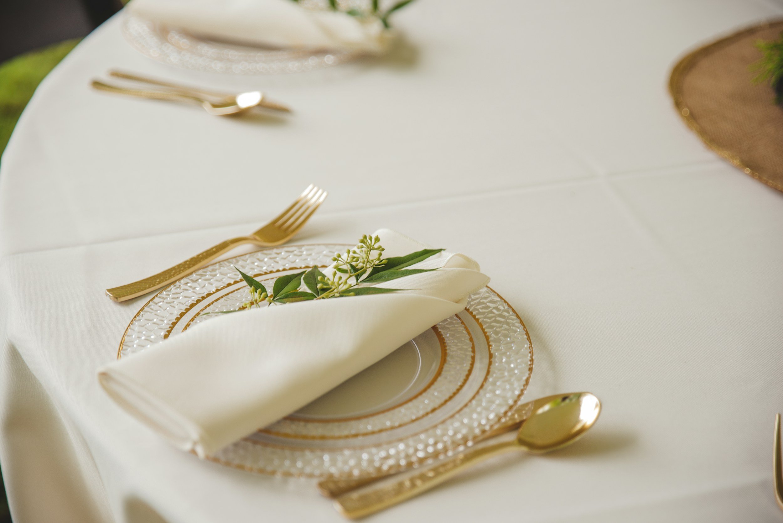 Elegant place setting on a white tablecloth with gold-rimmed plates, gold cutlery, a white napkin with a green sprig, and a decorative gold and brown placemat.