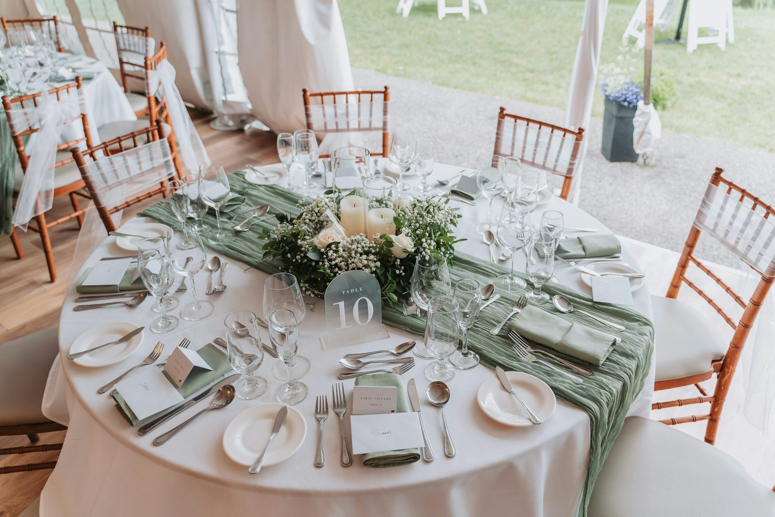 Round banquet table set for a wedding or event, decorated with green table runner, white candles, floral centerpiece with white roses and baby's breath, and multiple wine glasses, plates, and silverware, with place cards and a table number card reading '10'.