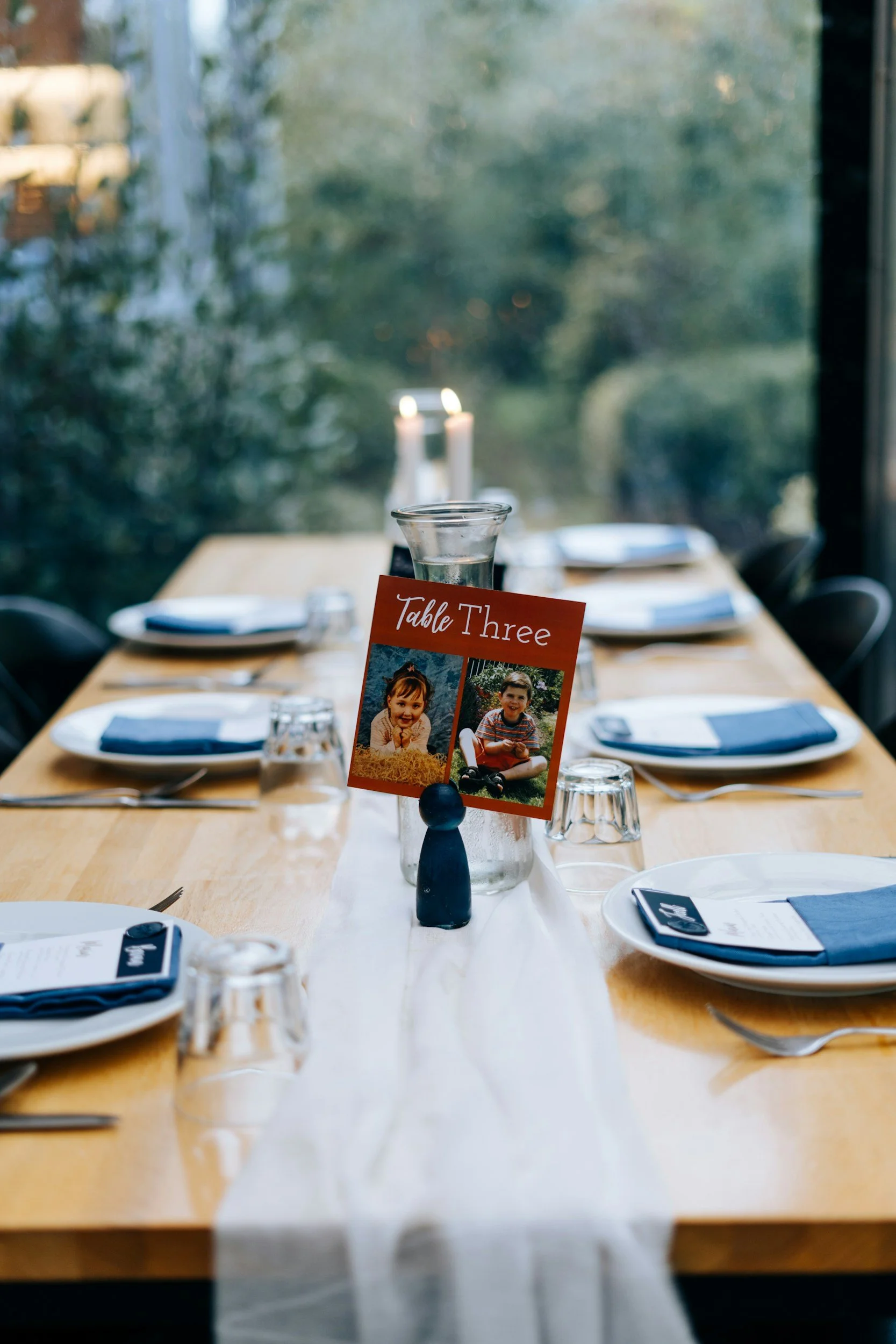 A dining table set with plates, glasses, cutlery, and folded napkins, with a sign labeled "Table Three" featuring photos of two children.