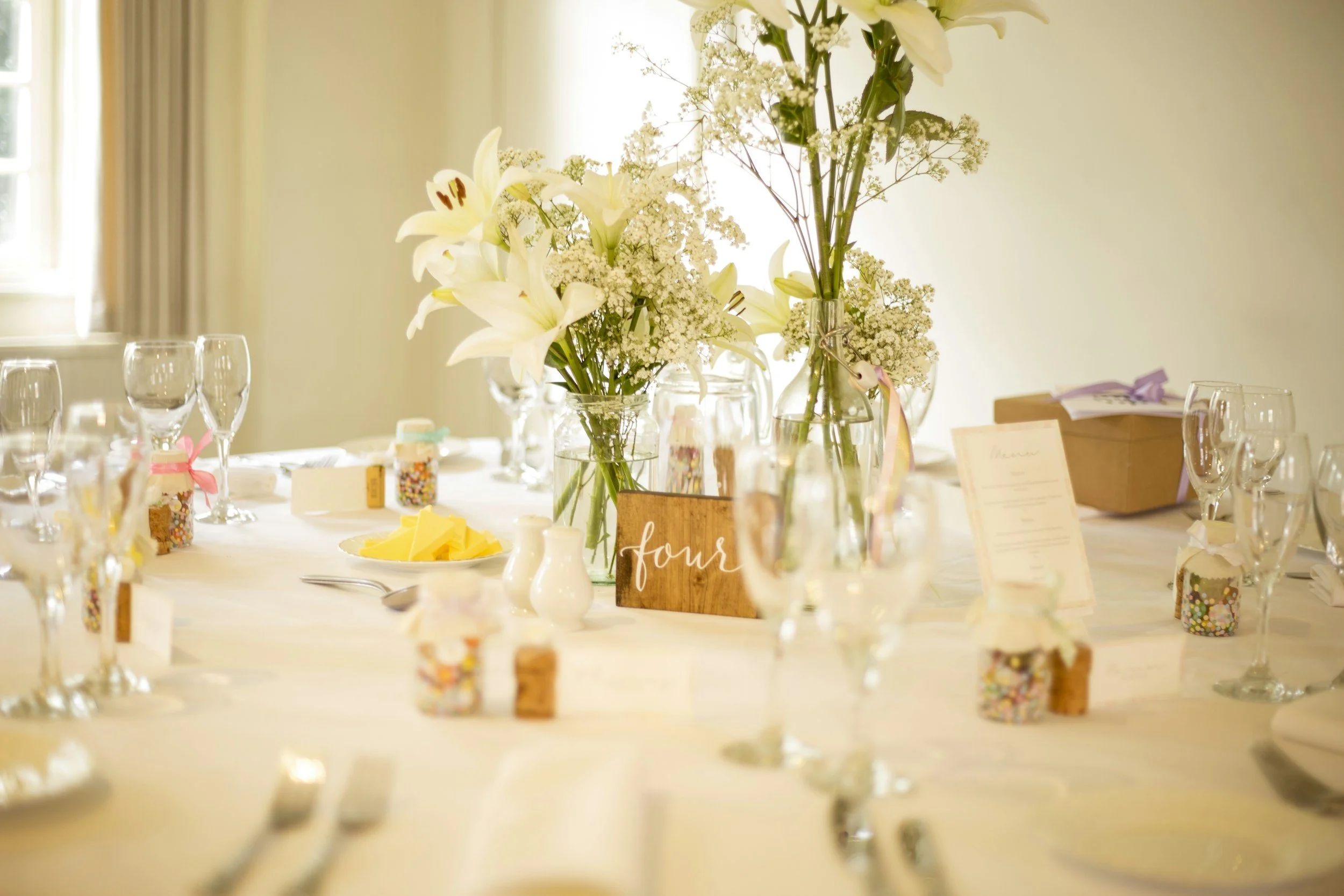 A table decorated with a floral centerpiece featuring white lilies and baby's breath, surrounded by empty wine glasses and small jars filled with colorful sprinkles, set for a celebration or event.