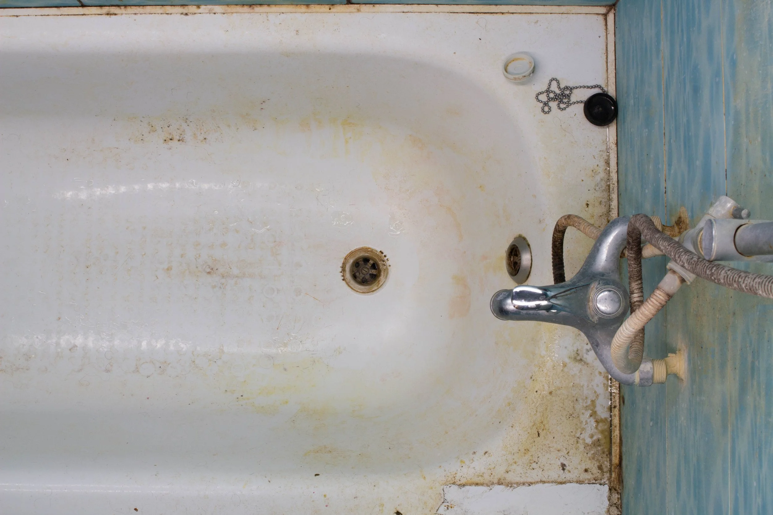 Old, dirty bathtub with rust stains, a faucet with attached hoses, a drain, and miscellaneous items including a chain and loose caps at the edge.