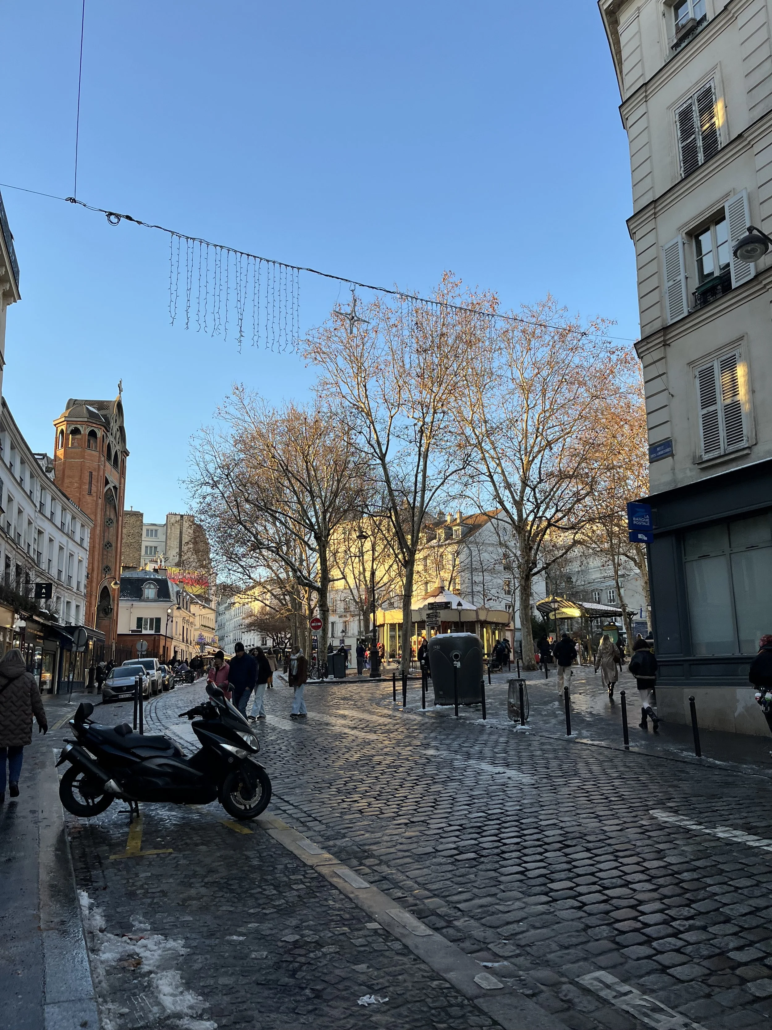 A cobblestone street in a European city with parked motorcycles, pedestrians, leafless trees, and historic buildings under a clear blue sky.