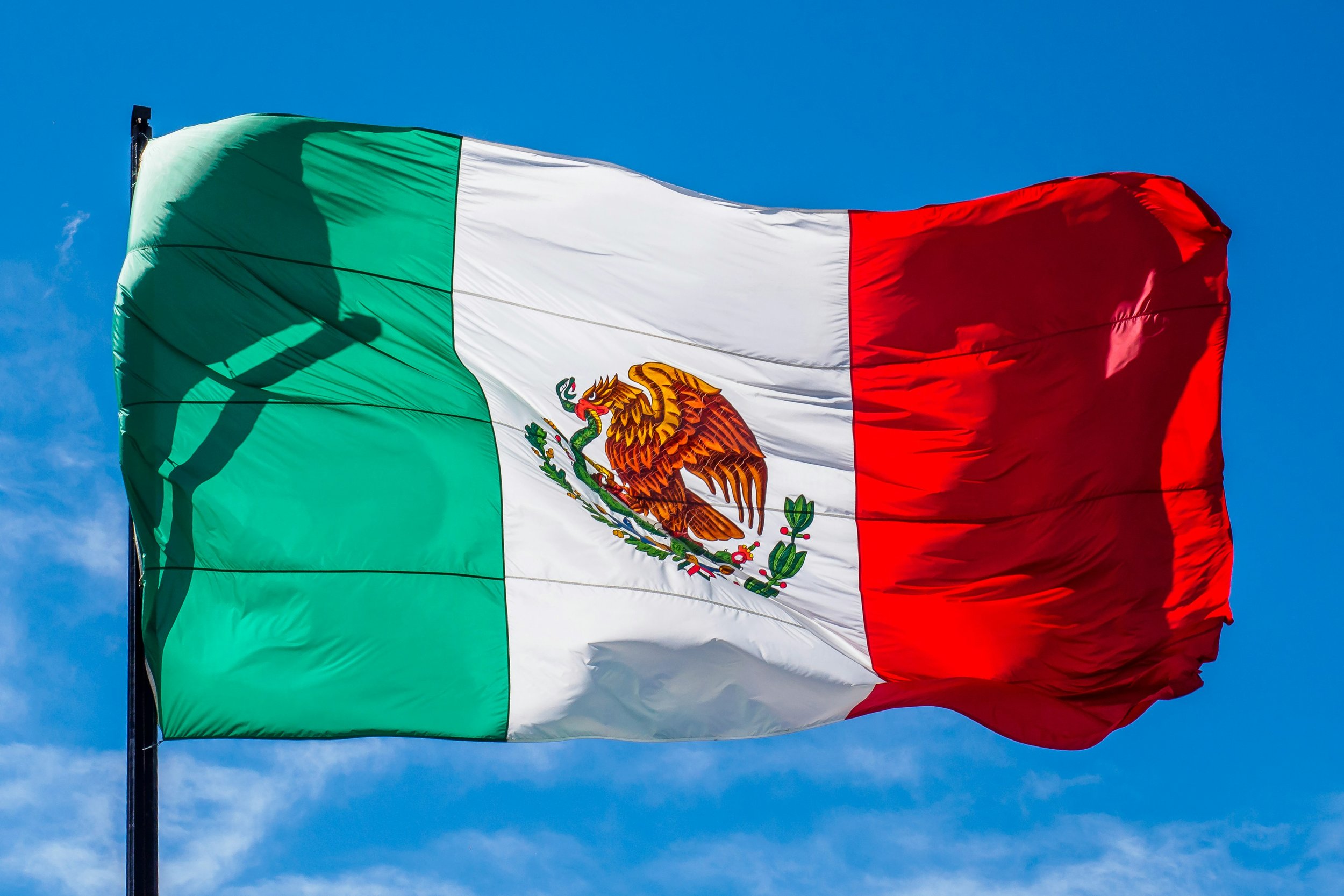 Mexican flag waving in the wind against a blue sky, featuring green, white, and red vertical stripes with a coat of arms of an eagle eating a snake on a cactus in the center of the white stripe.