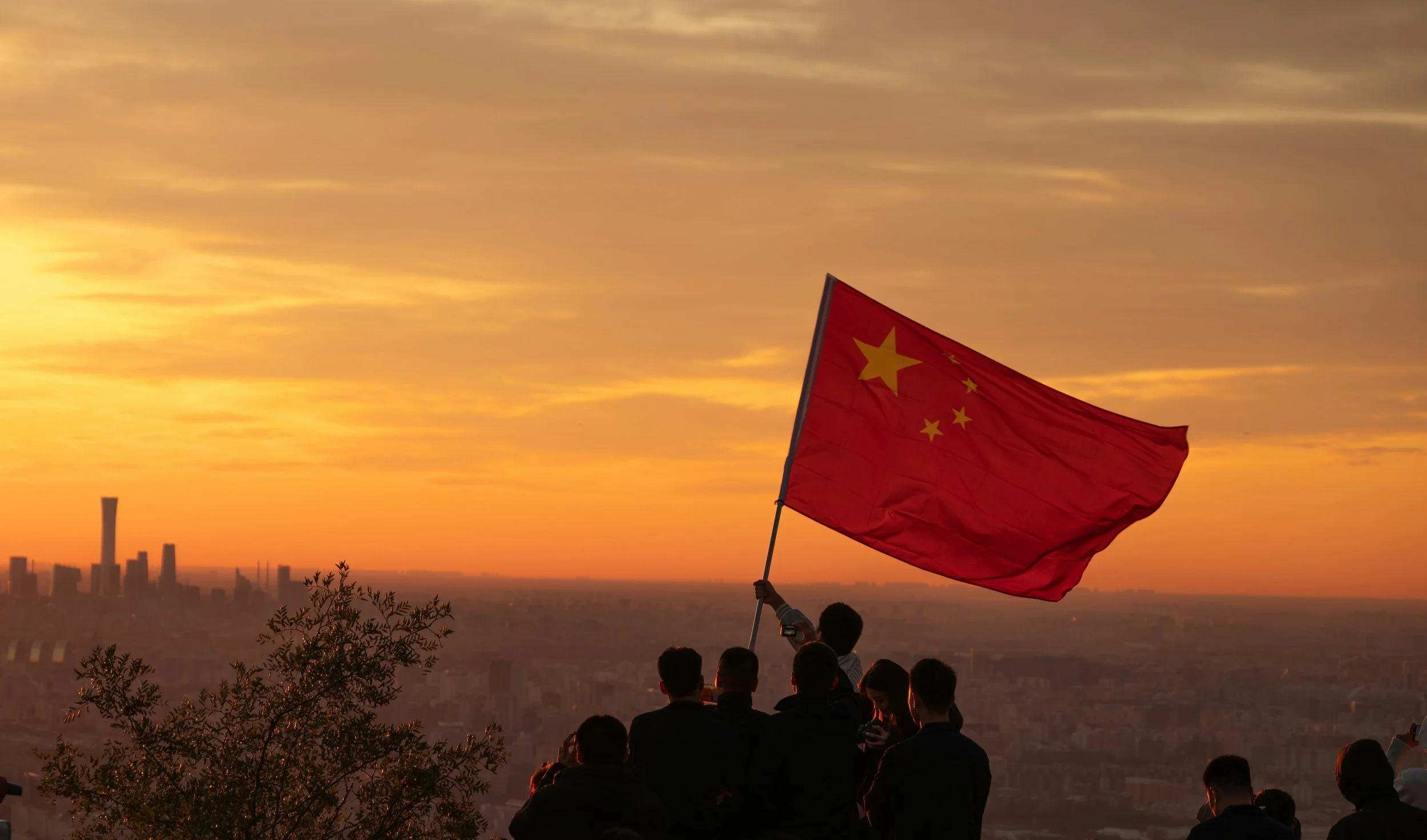 A group of people on a hilltop watching a sunset, with one person holding a large Chinese flag.