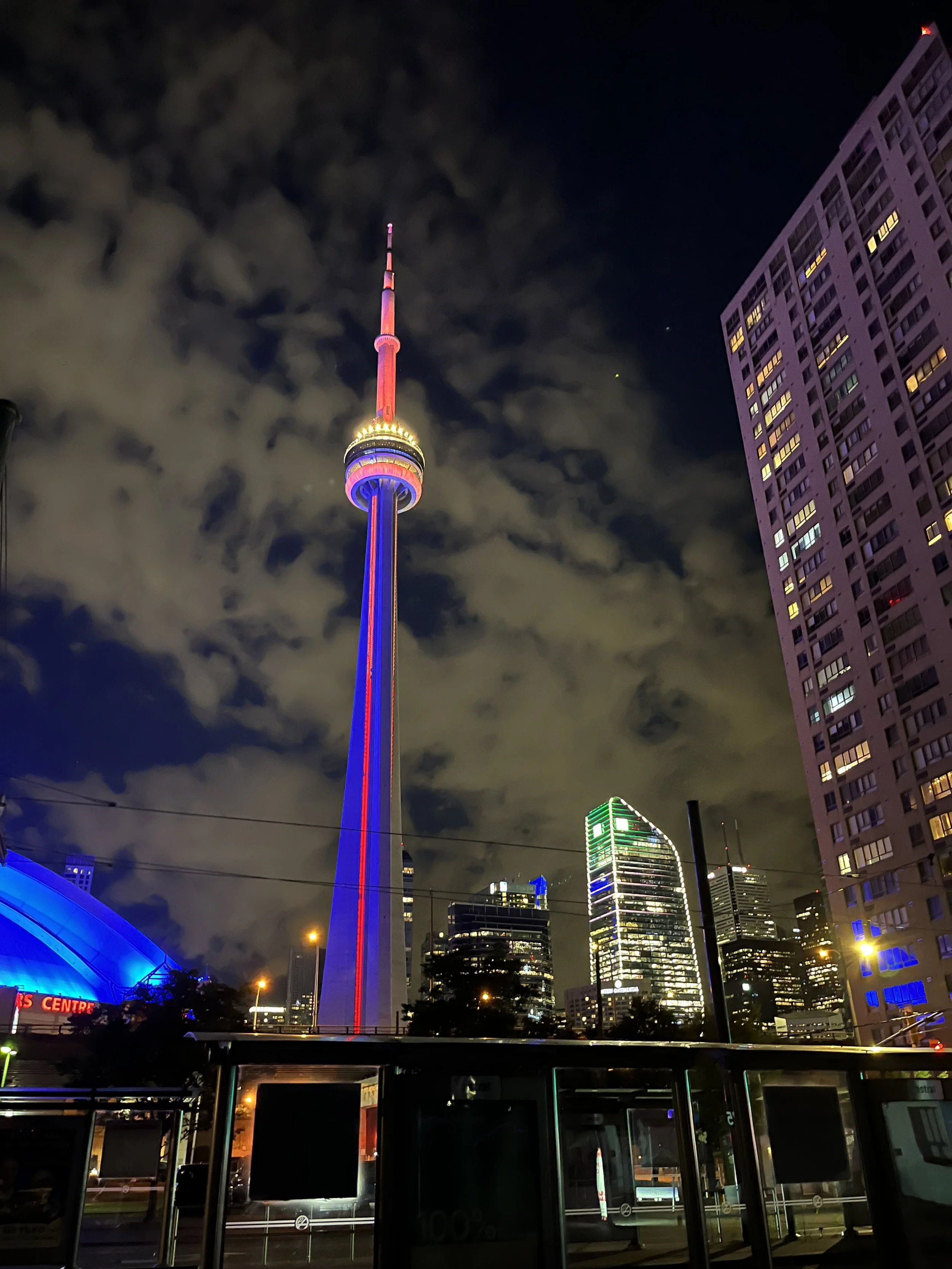 Night view of Toronto skyline featuring the CN Tower illuminated in red and blue lights, with surrounding high-rise buildings and cloudy sky.