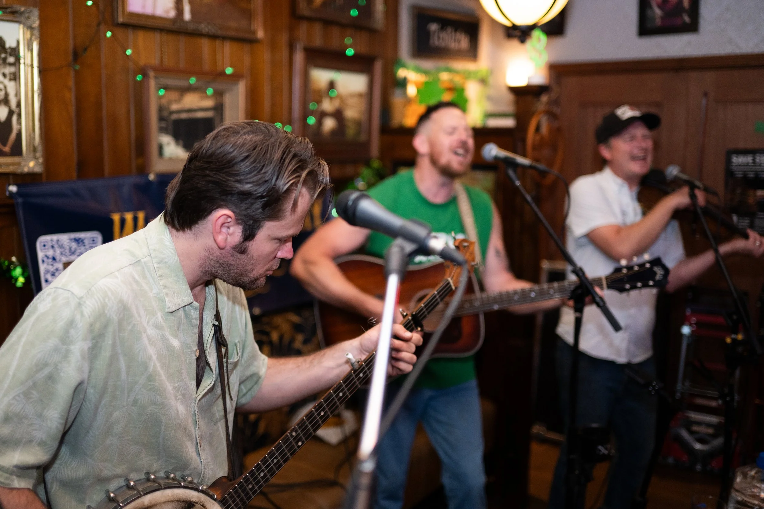 Three musicians performing with guitars in a cozy, decorated indoor setting, with two of them singing into microphones and one playing a banjo.