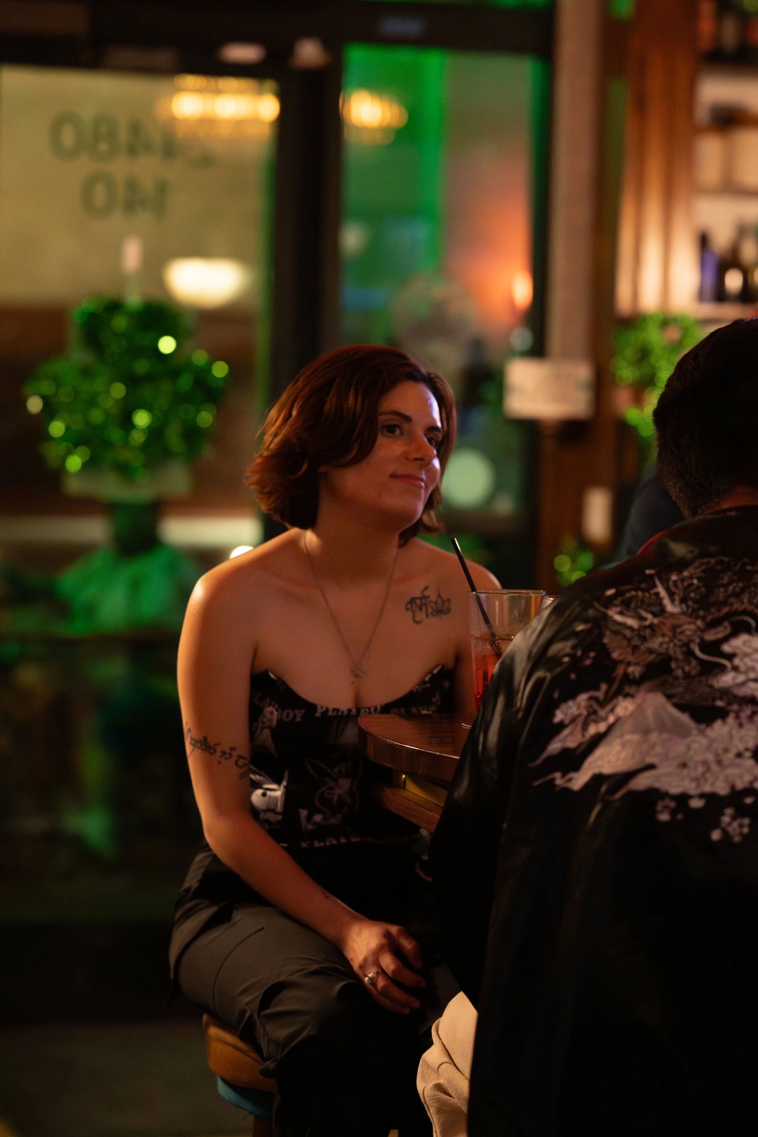 A woman with reddish-brown hair sitting at a bar table with a drink that has a straw, engaged in conversation with a man wearing a patterned shirt, with a bartender and greenery in the background.