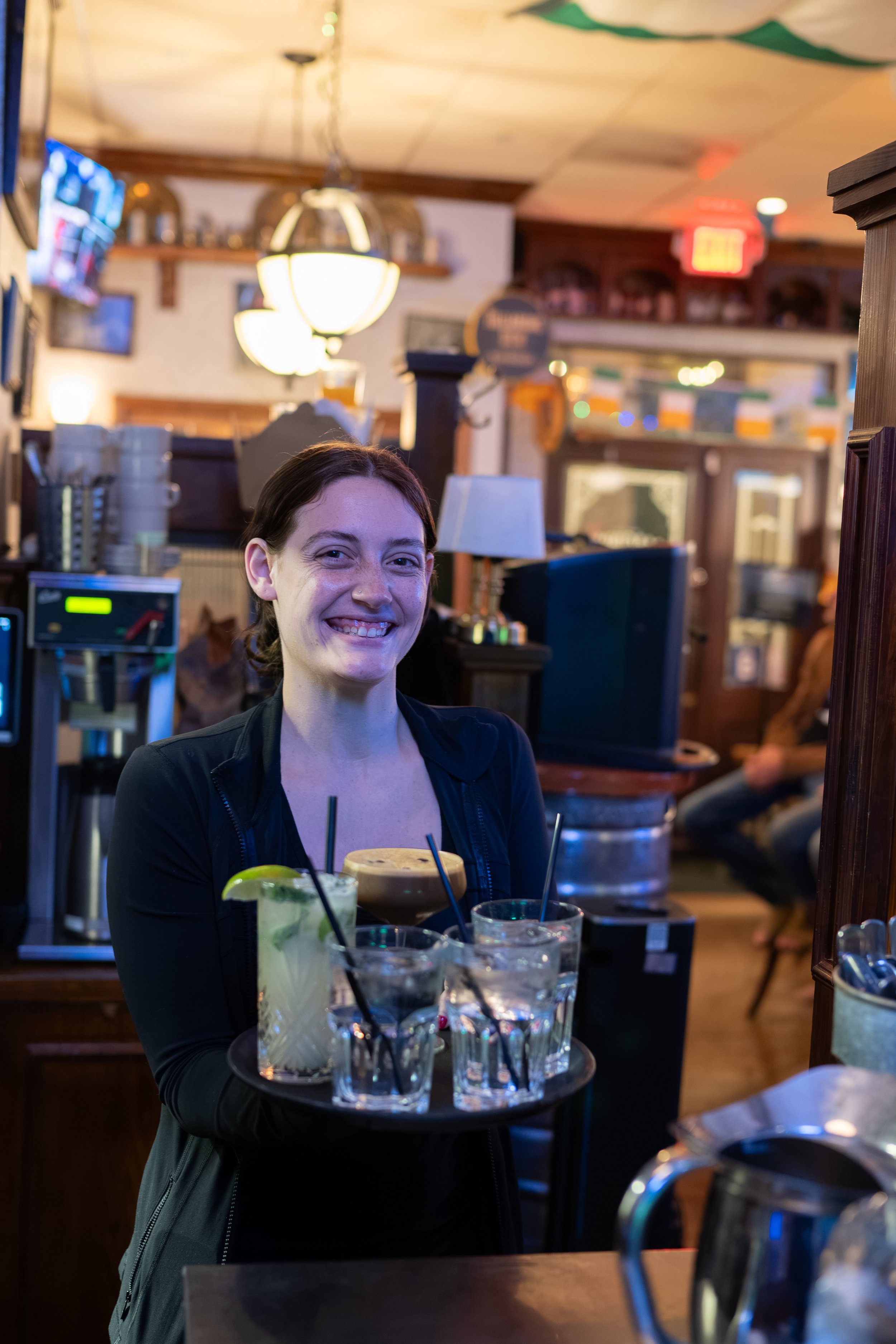 A woman smiling while holding a tray with four drinks inside a restaurant or bar, with wooden decor and warm lighting in the background.