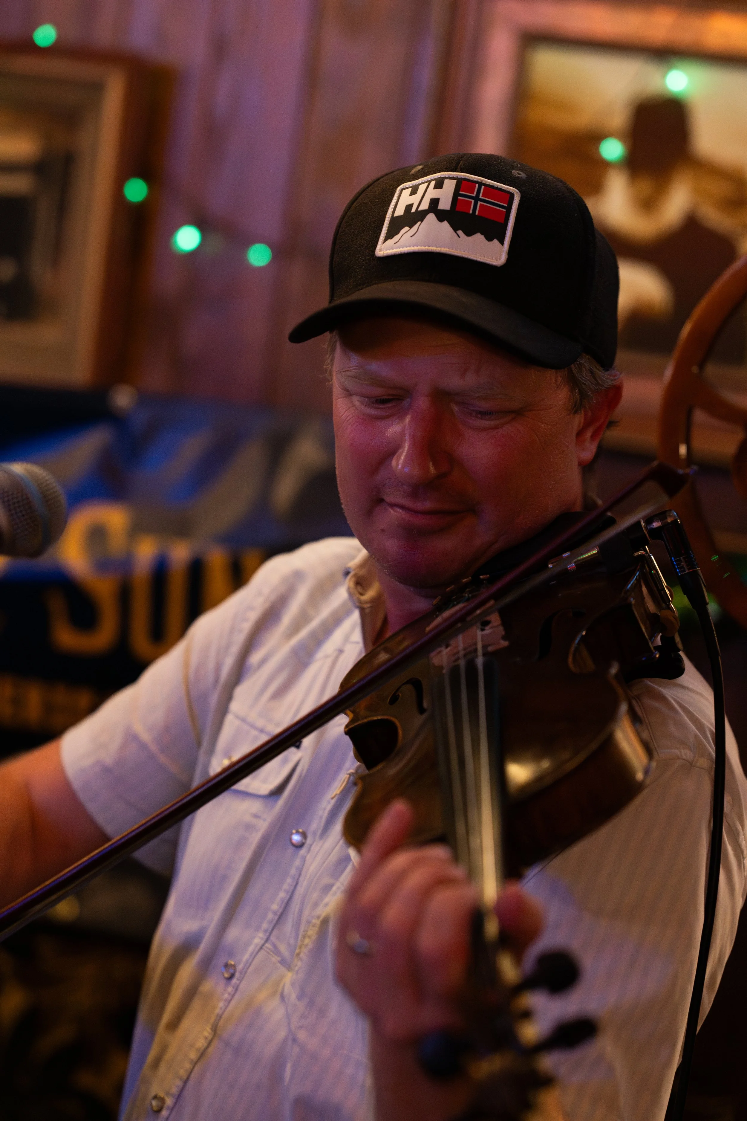 A man playing a violin on stage, wearing a black cap with a logo, in a cozy setting with fairy lights and wooden decor.