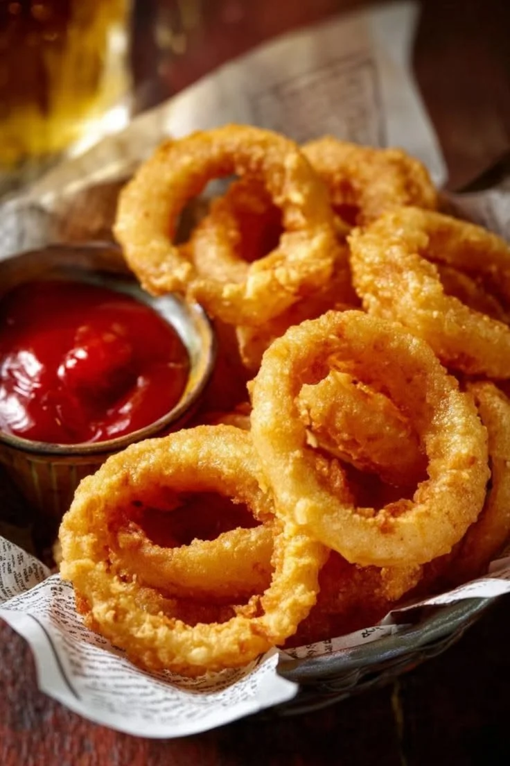 Golden fried onion rings served with ketchup in a small metal cup.