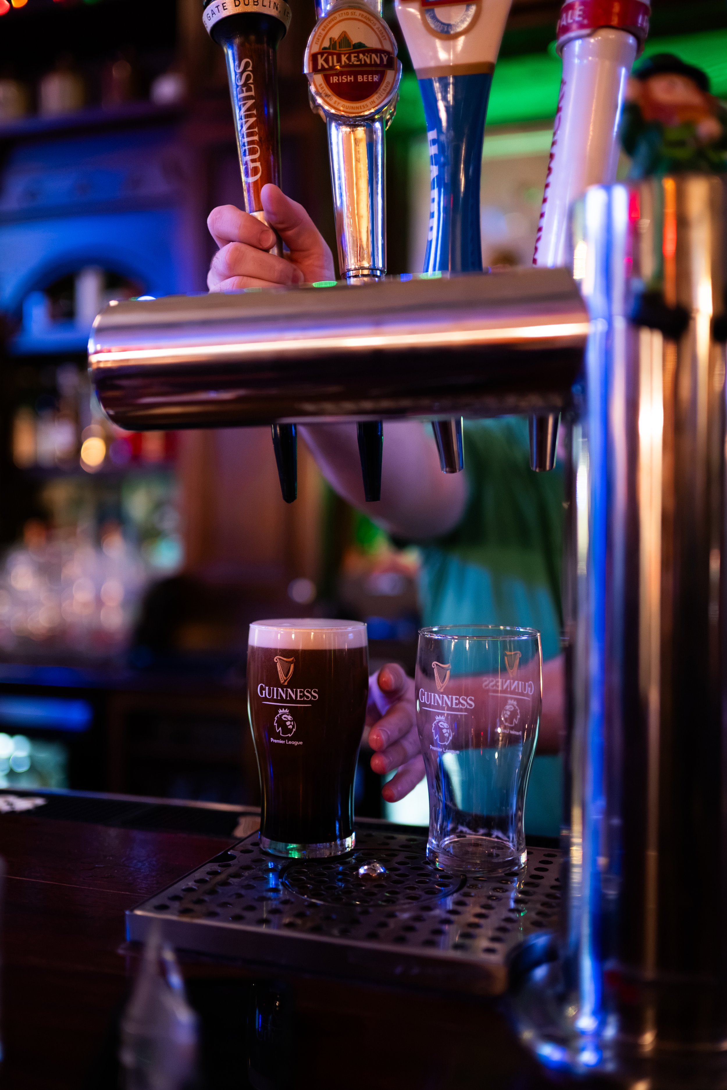 A bartender's hand is placing Guinness beer taps on a bar counter; two Guinness glasses are on the bar, one filled with dark beer and one empty, both with Guinness branding, in a lively pub setting with colorful lighting.