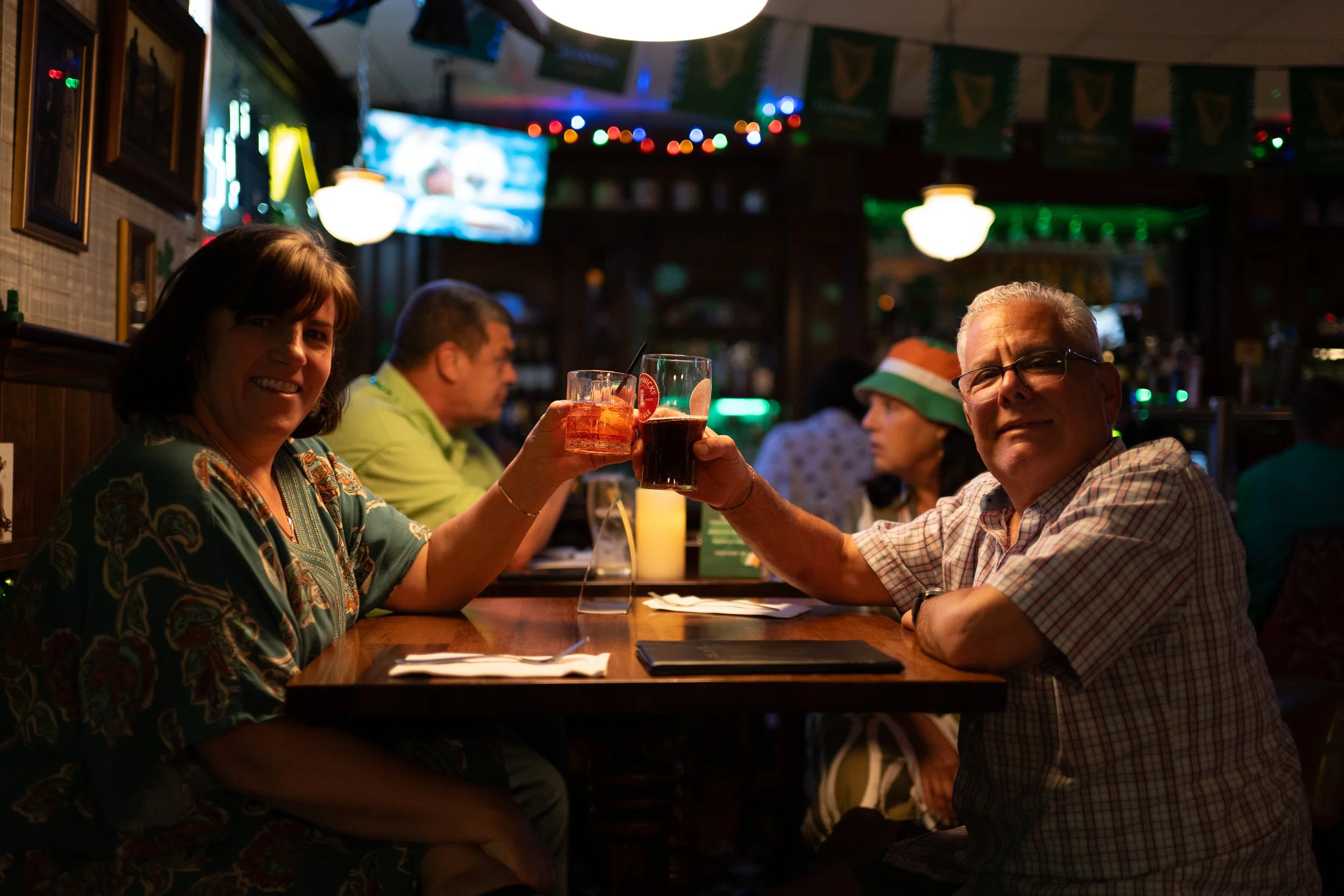 Two older adults sitting at a bar, smiling, raising their drinks for a toast, with other patrons in the background in a dimly lit pub decorated with Irish flags and string lights.