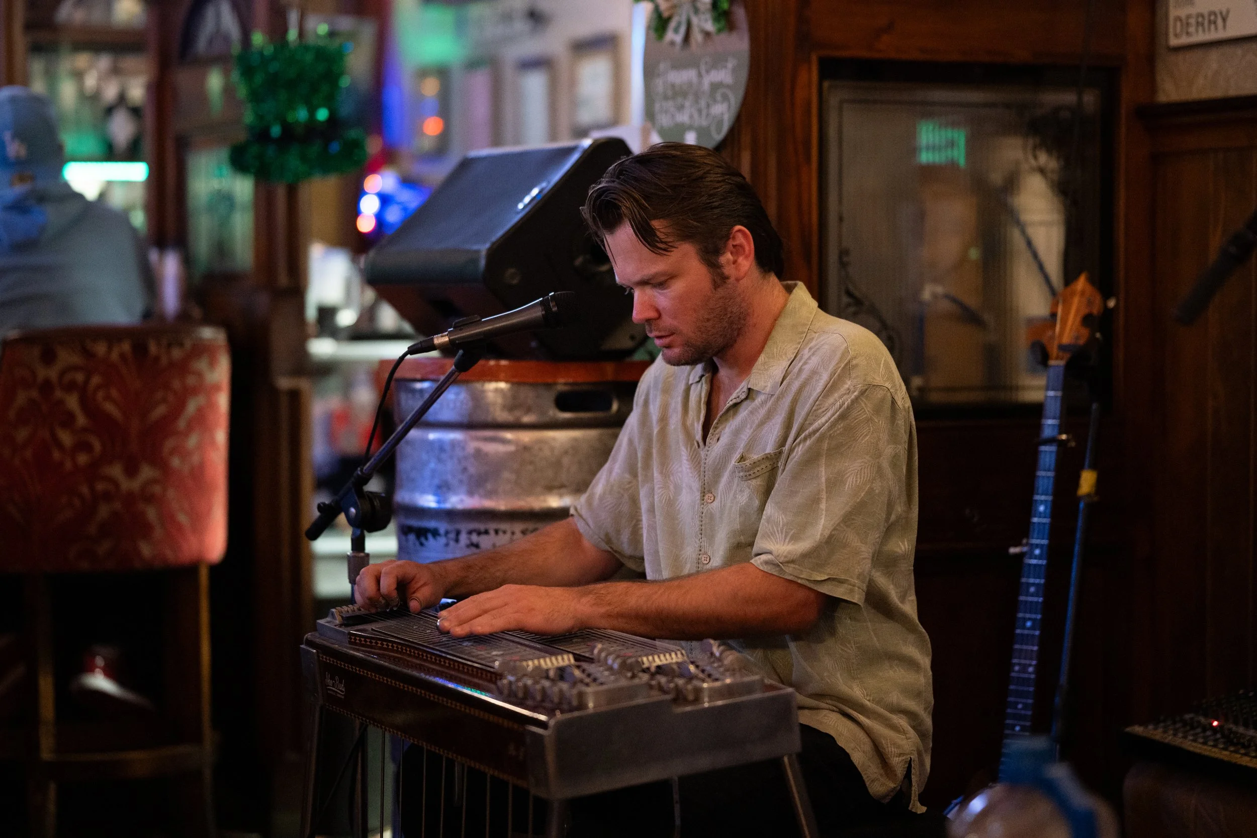 A man playing a lap steel guitar at a bar with wooden walls, microphone, and musical instruments in the background.