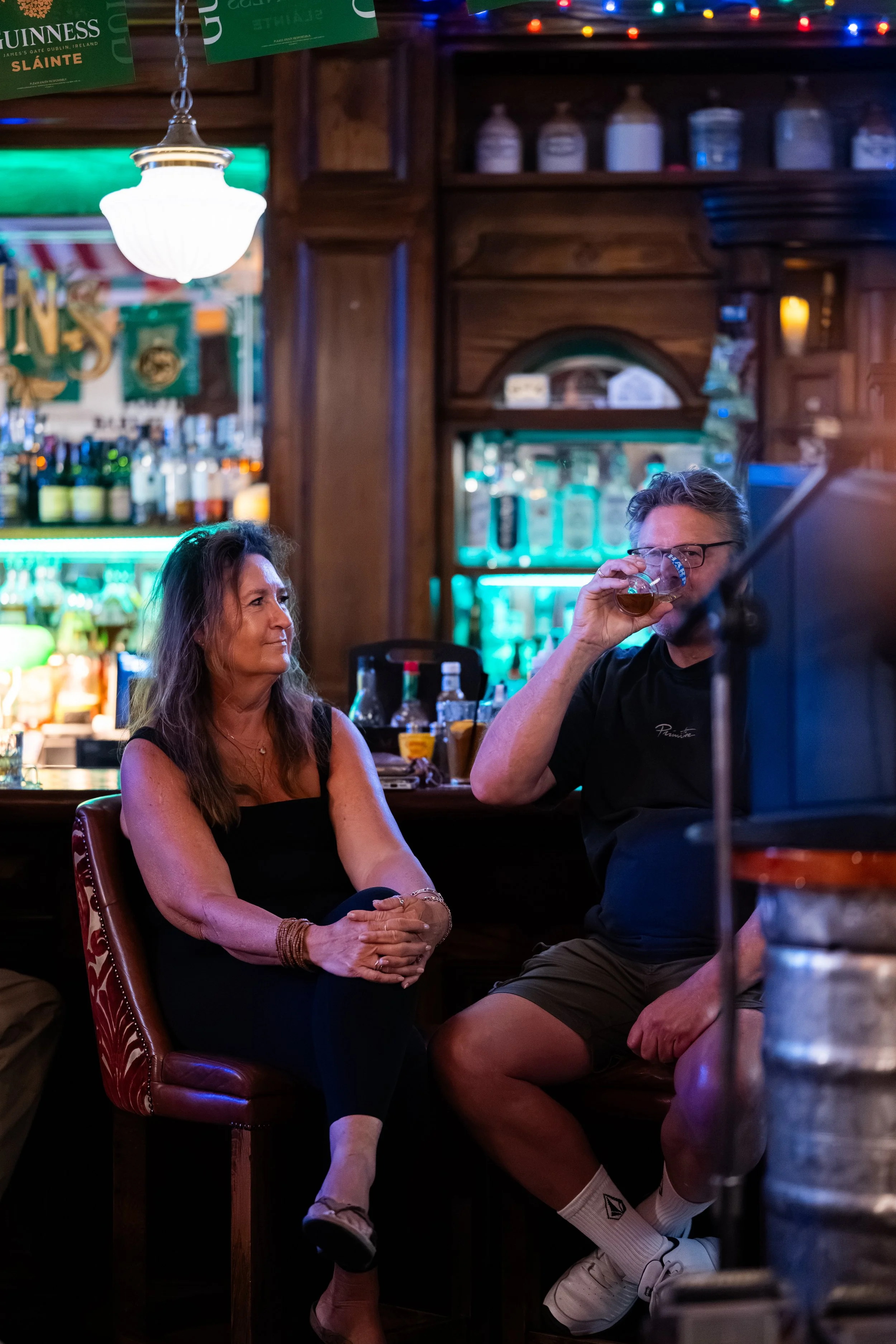 A woman with long brown hair sitting at a bar and looking at a man with short hair, glasses, and a beard, who is drinking from a glass. The bar has wooden shelves with bottles, jars, and colorful Christmas lights overhead.