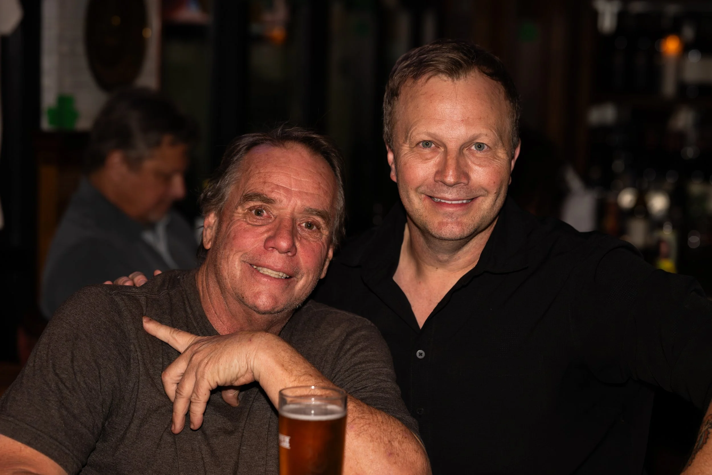 Two men smiling, one with his hand on the shoulder of the other, at a social gathering or bar. A glass of beer is in front of one of them, and a man is blurred in the background.