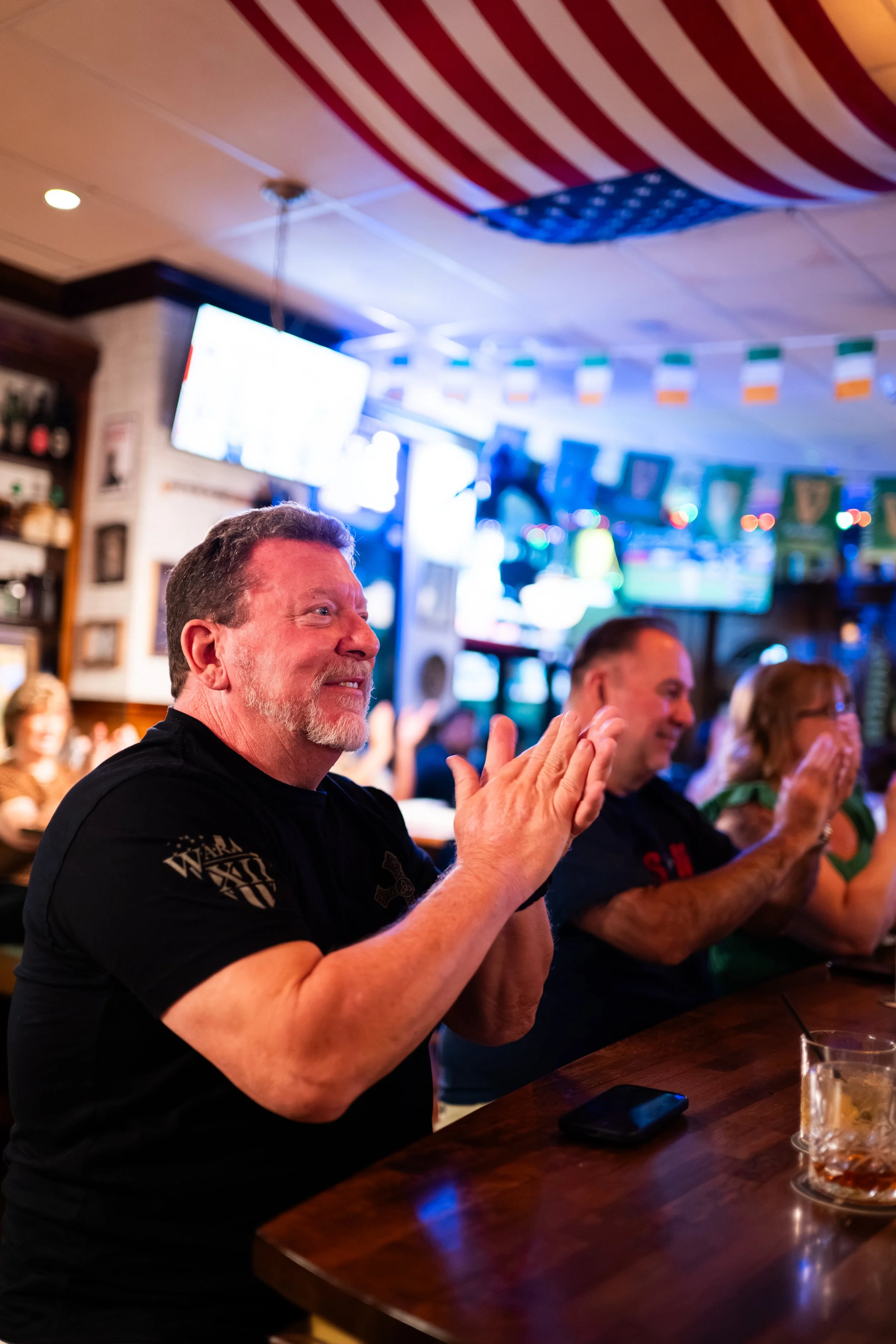 People, including a man with a beard and a black shirt, clapping and enjoying themselves in a bar or pub with American flags hanging from the ceiling and television screens in the background.