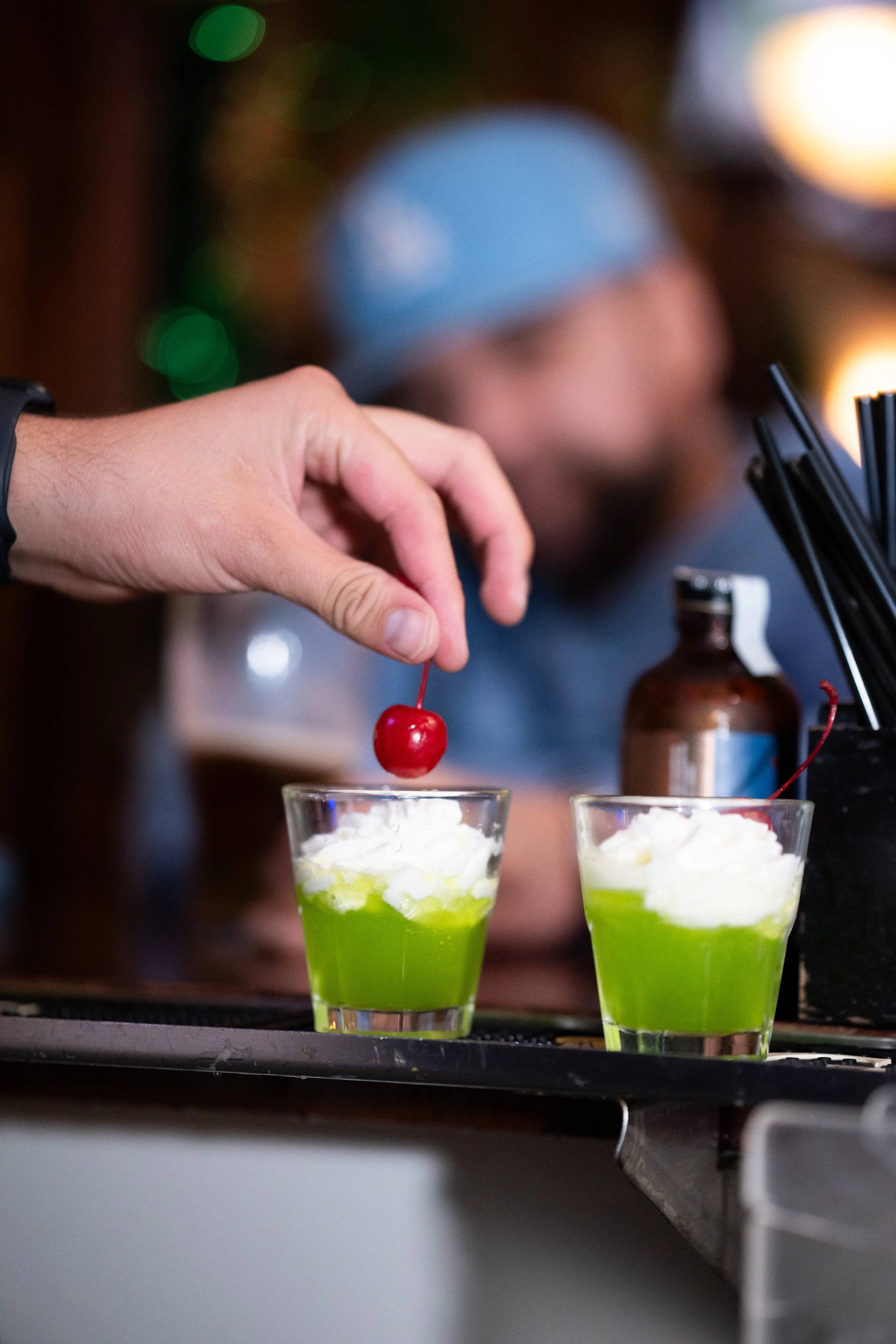 A person is garnishing two green dessert drinks with whipped cream and cherries, with one cherry being placed on top.