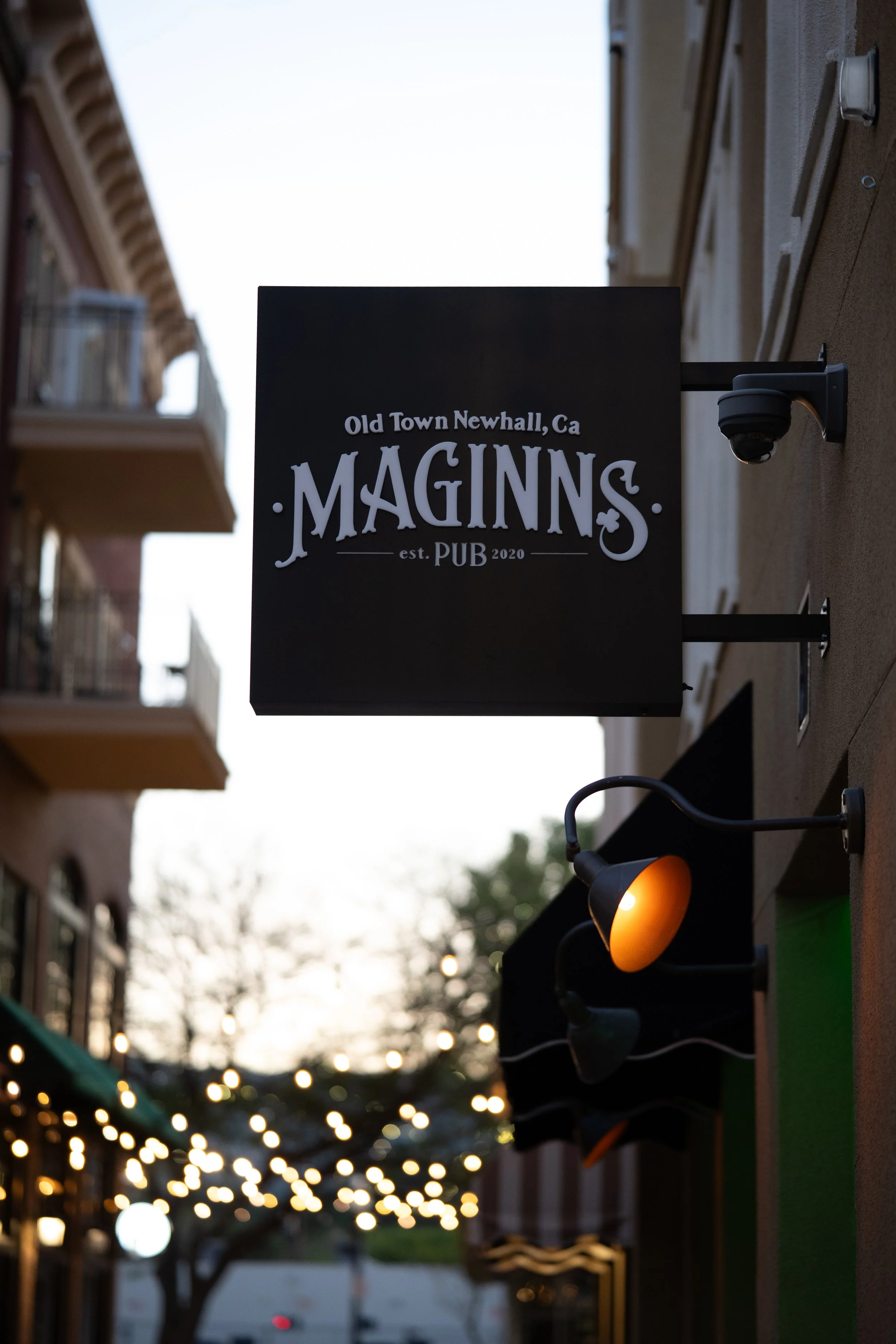 Sign for Old Town Newhall, California, Maginns Pub, established 2020 with outdoor string lights and security camera.