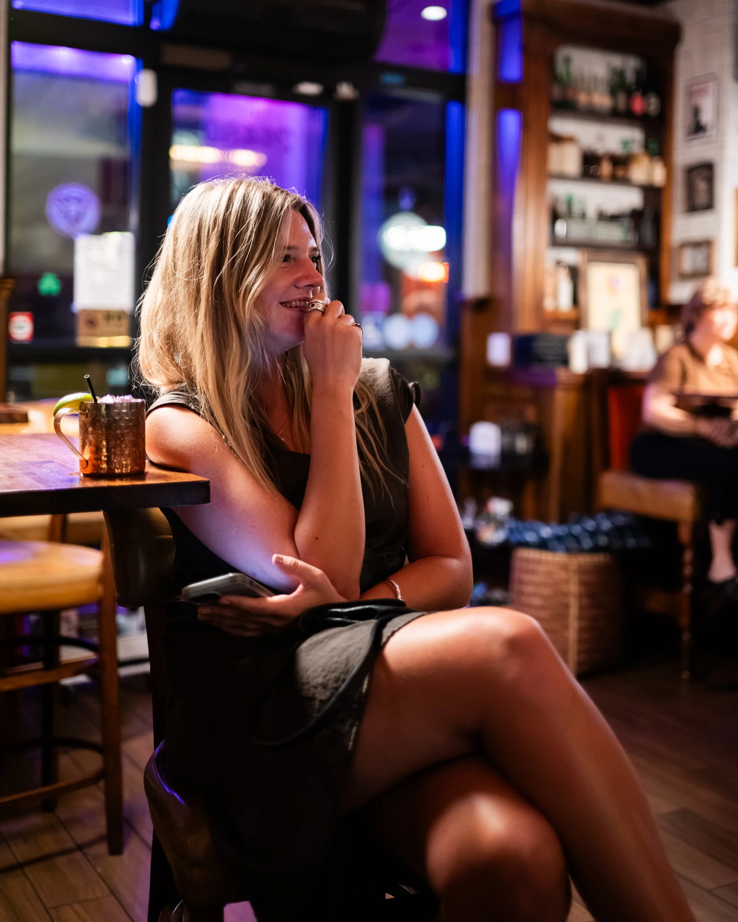 A woman with long blonde hair, wearing a black top and shorts, sitting at a wooden table in a dimly lit restaurant or bar, smiling with her hand near her mouth. In front of her is a mug with a lime wedge and a straw. The background shows shelves with