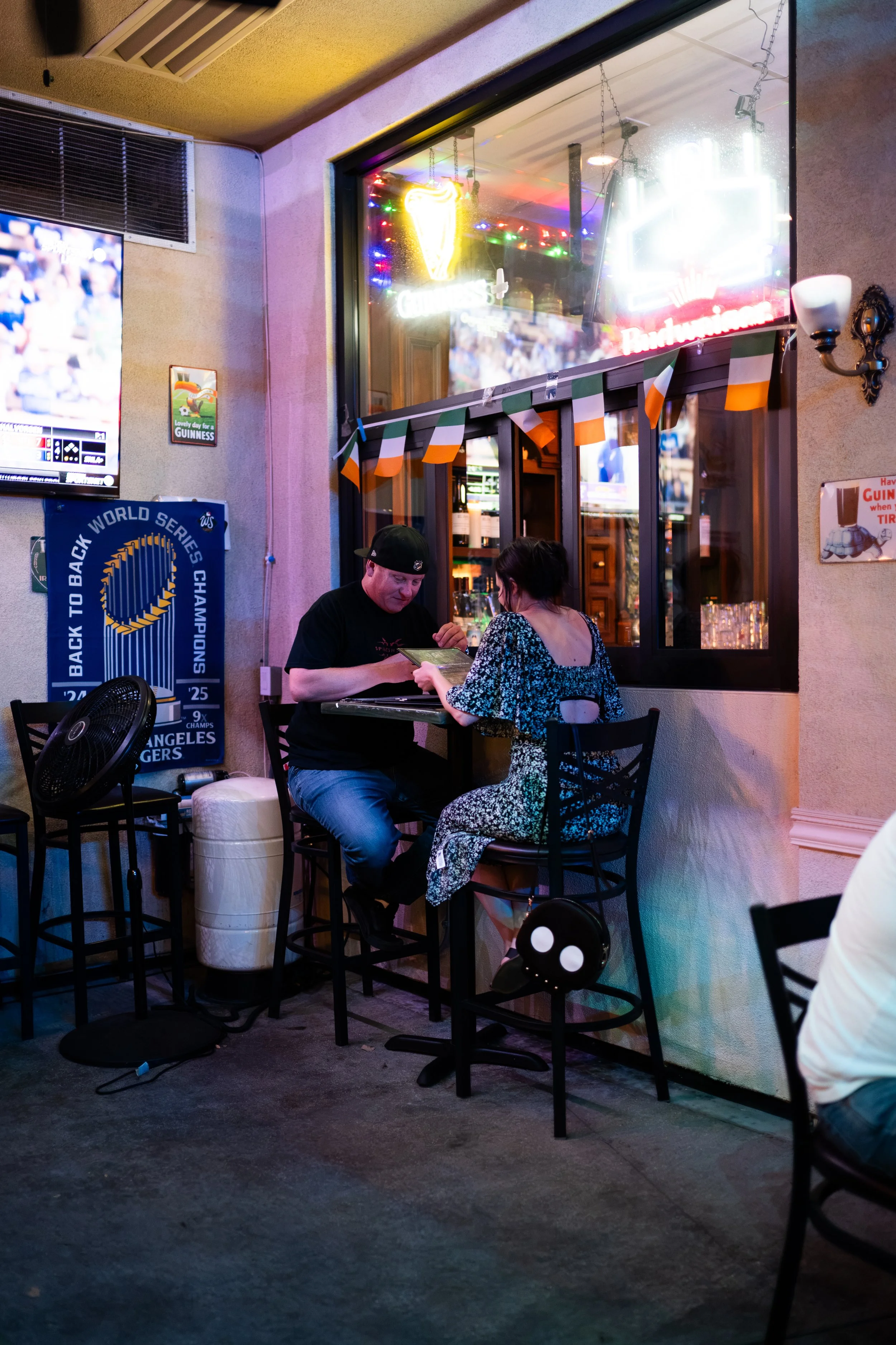 Two people sitting at a high table inside a bar or restaurant, engaged in conversation, with neon signs, a television, and decorations such as flags and banners visible in the background.