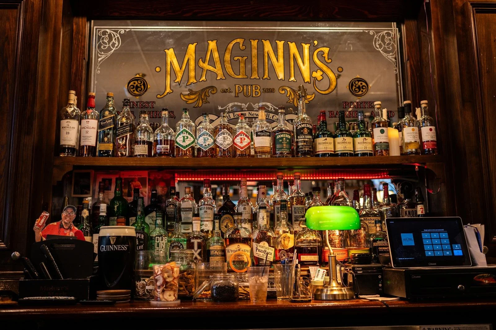 Bar with various bottles of whiskey and liquor on shelves, a bartender behind the counter, and a large sign reading "Maginn's Pub 2020" above the shelves.