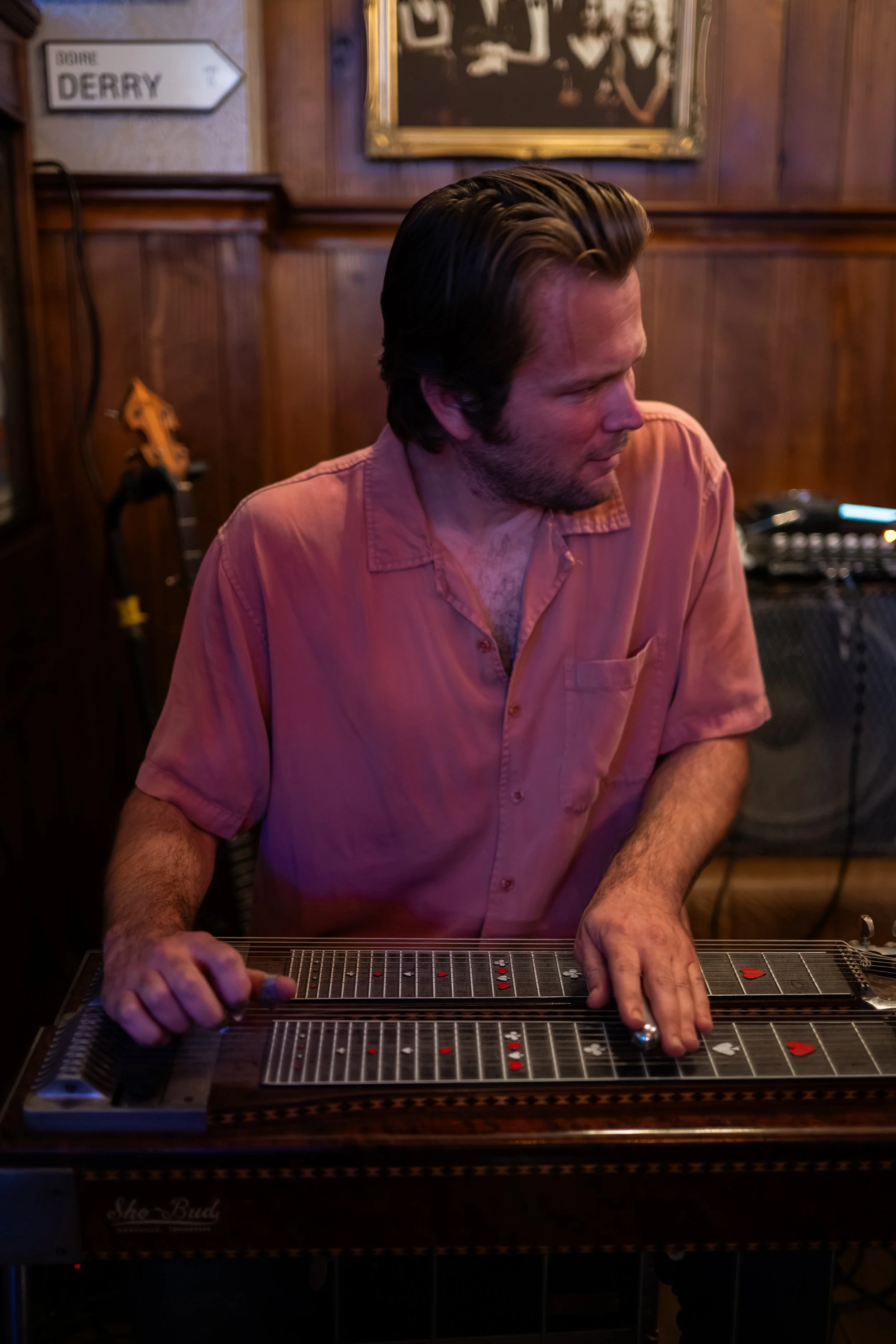 A man with brown hair and a beard, wearing a pink shirt, is playing a steel guitar in a wooden-paneled room with musical equipment in the background.