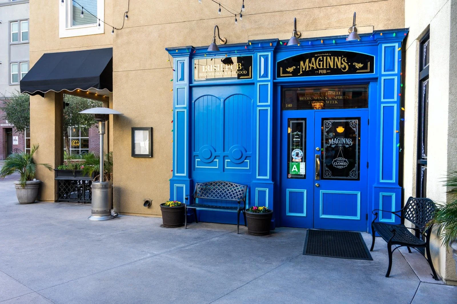 The entrance of Maginn's pub is painted bright blue with a decorative facade. There are potted plants and benches outside, and a black awning extends over an adjacent sidewalk area. String lights are hung above the entrance.
