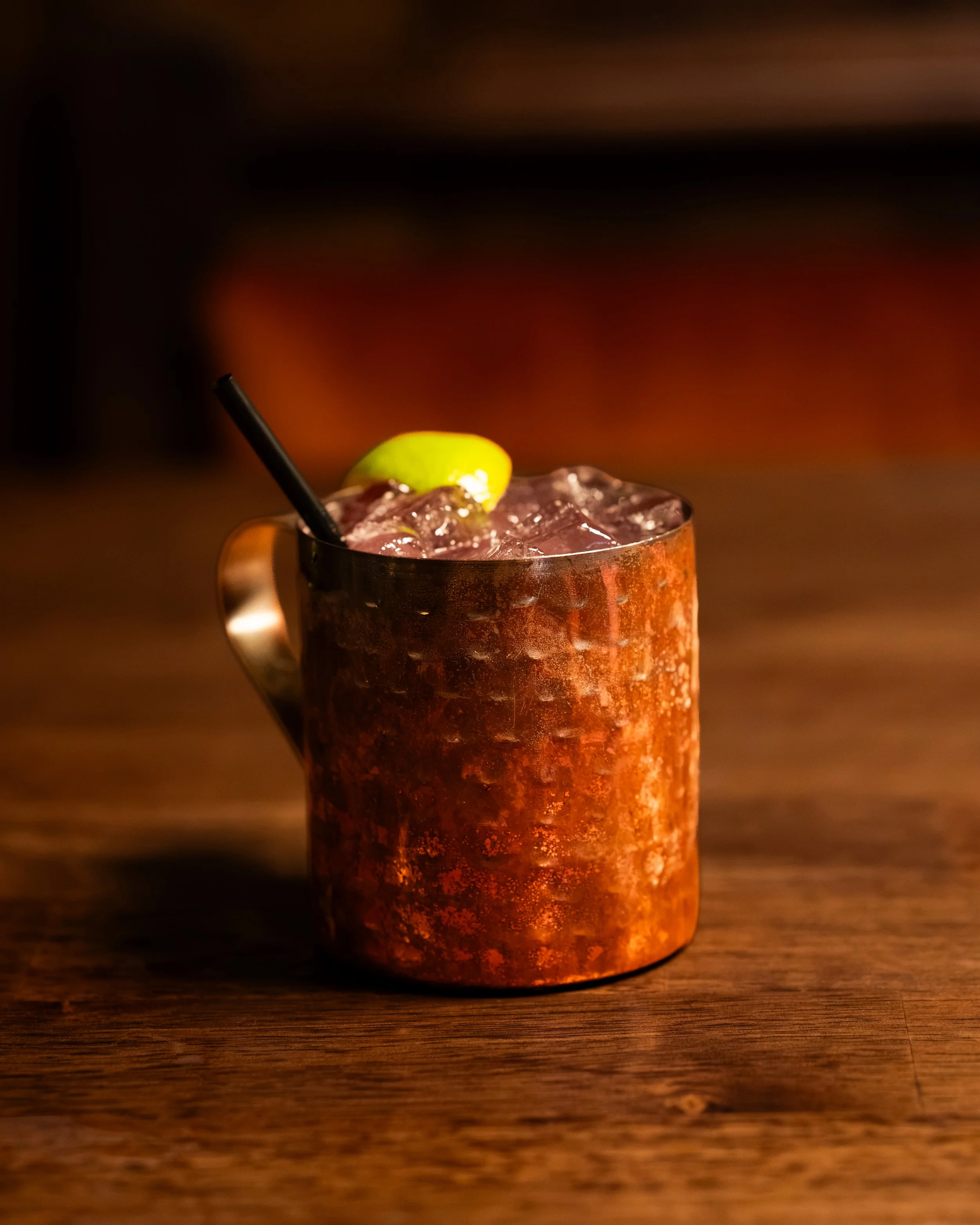 A copper mug filled with a dark beverage, ice cubes, garnished with a lime wedge and a black straw, placed on a wooden surface with a dark, blurred background.