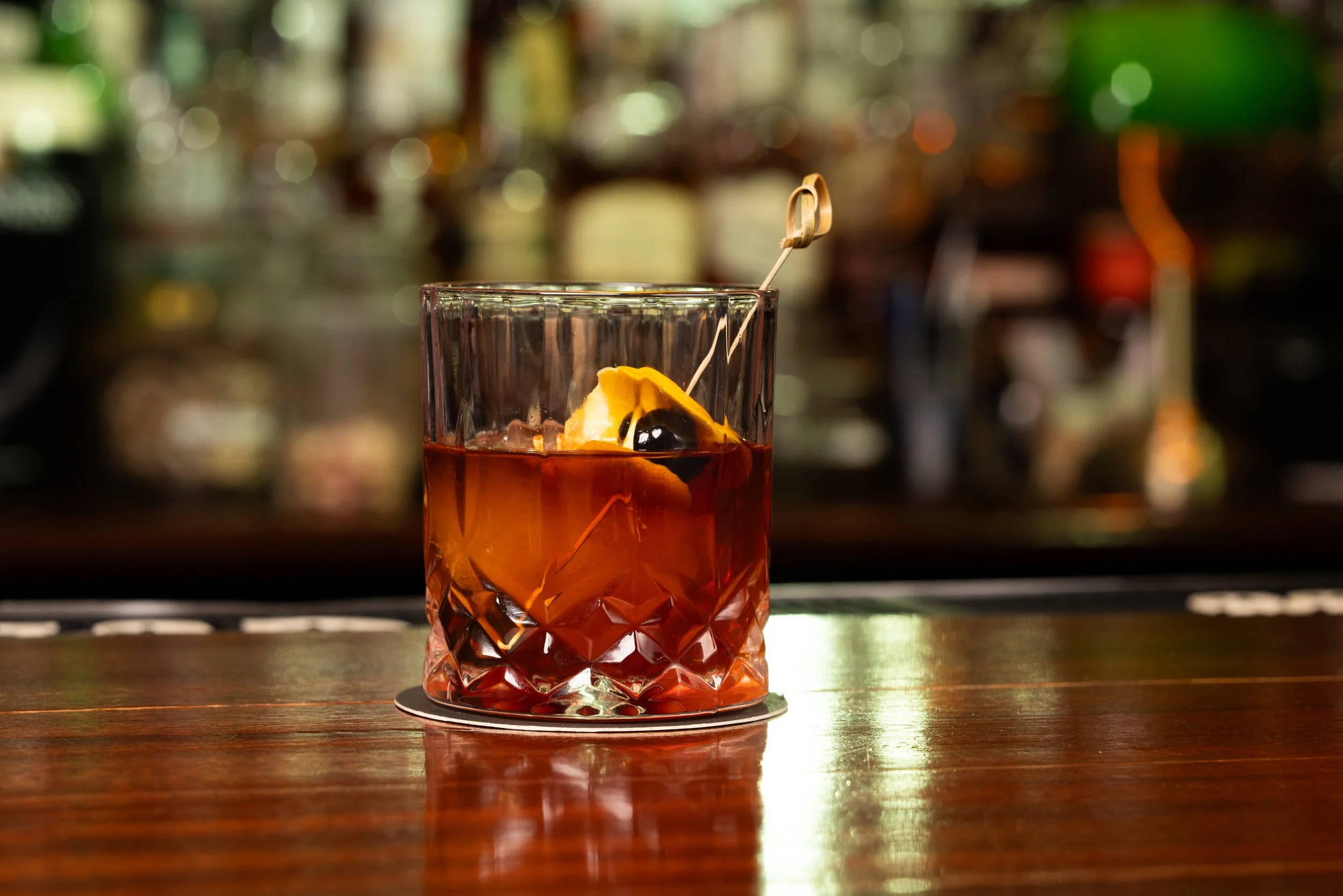 A glass of amber-colored cocktail with a lemon wedge and a cherry, garnished with a cocktail pick, on a bar counter.