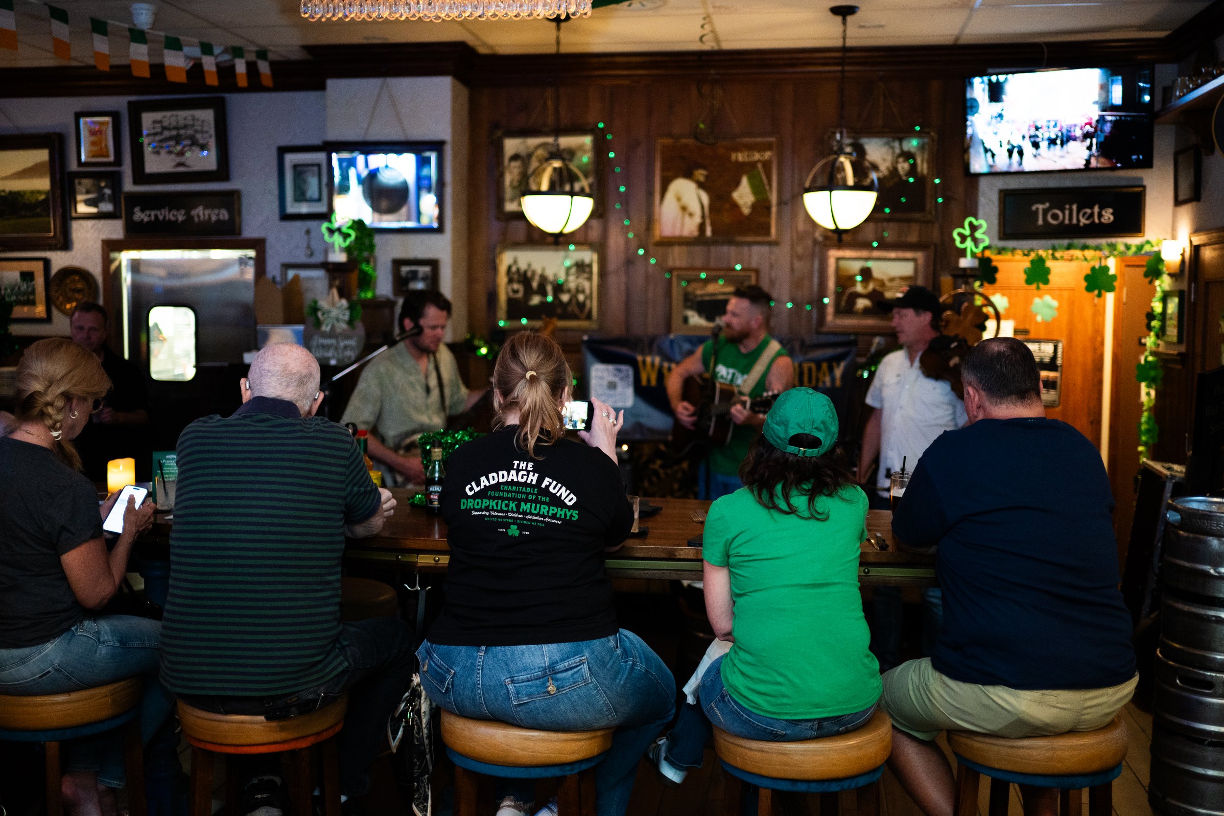 People watching a St. Patrick's Day celebration at a bar, with musicians playing guitar and singing, festive green decorations, and some patrons taking photos or using phones.