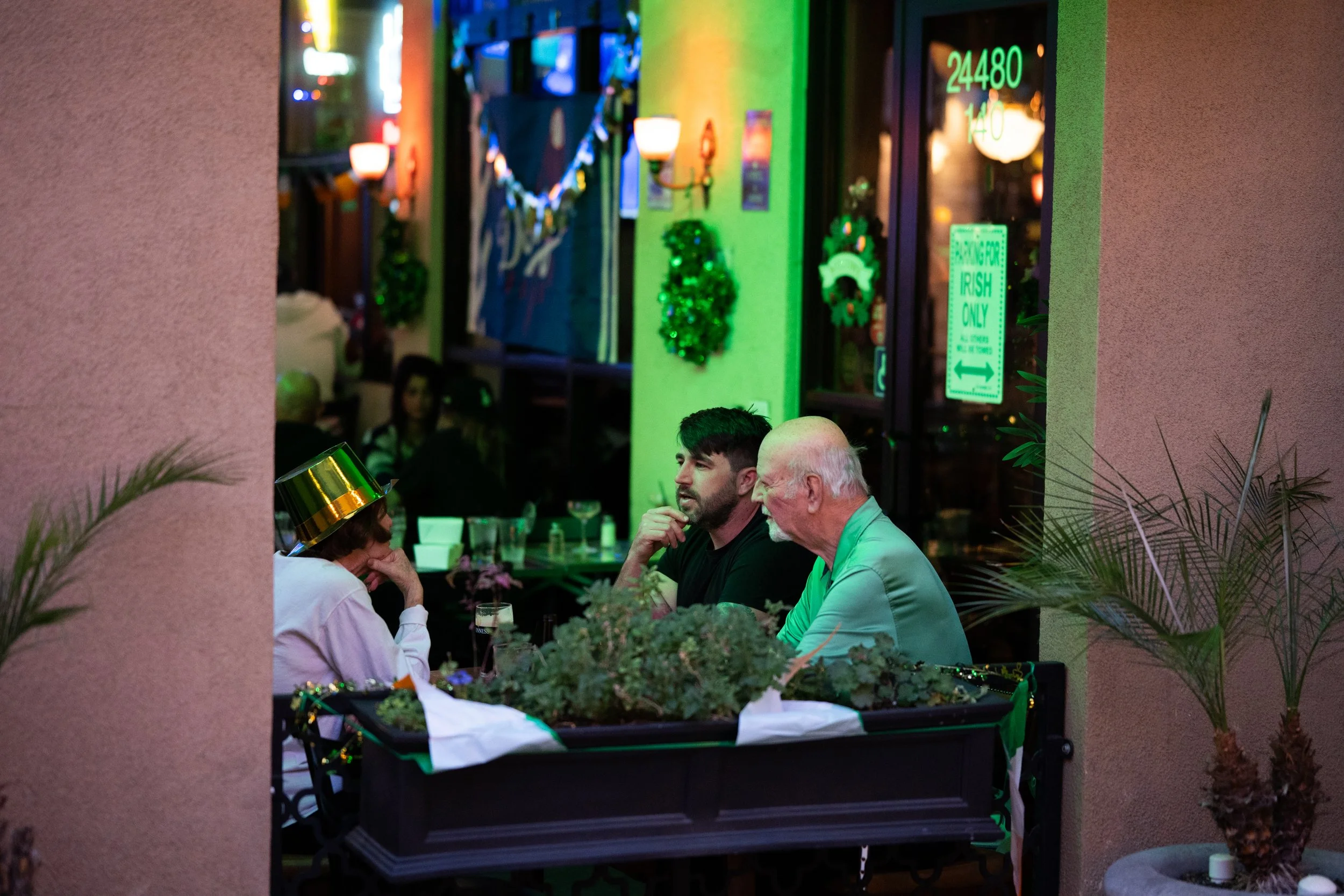 People celebrating St. Patrick's Day at a restaurant, including one wearing a shiny green and gold hat, with green decorations and neon signs visible in the background.