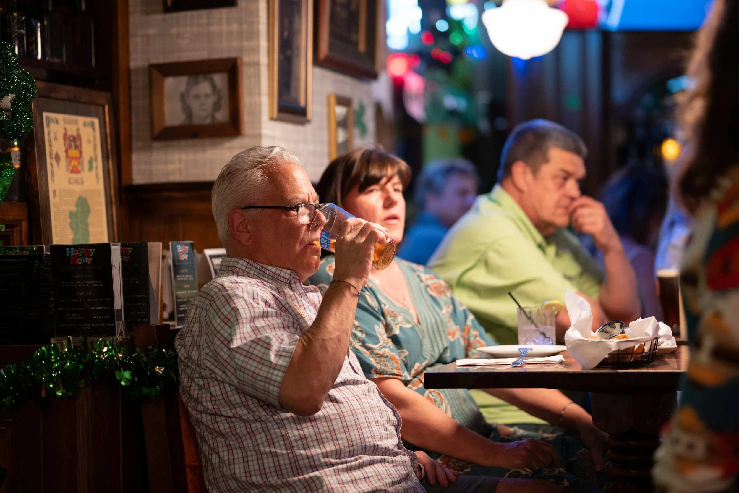 People sitting at a table in a decorated restaurant, with one man drinking a beer, a woman, and other individuals in the background, with holiday decorations and dim lighting.