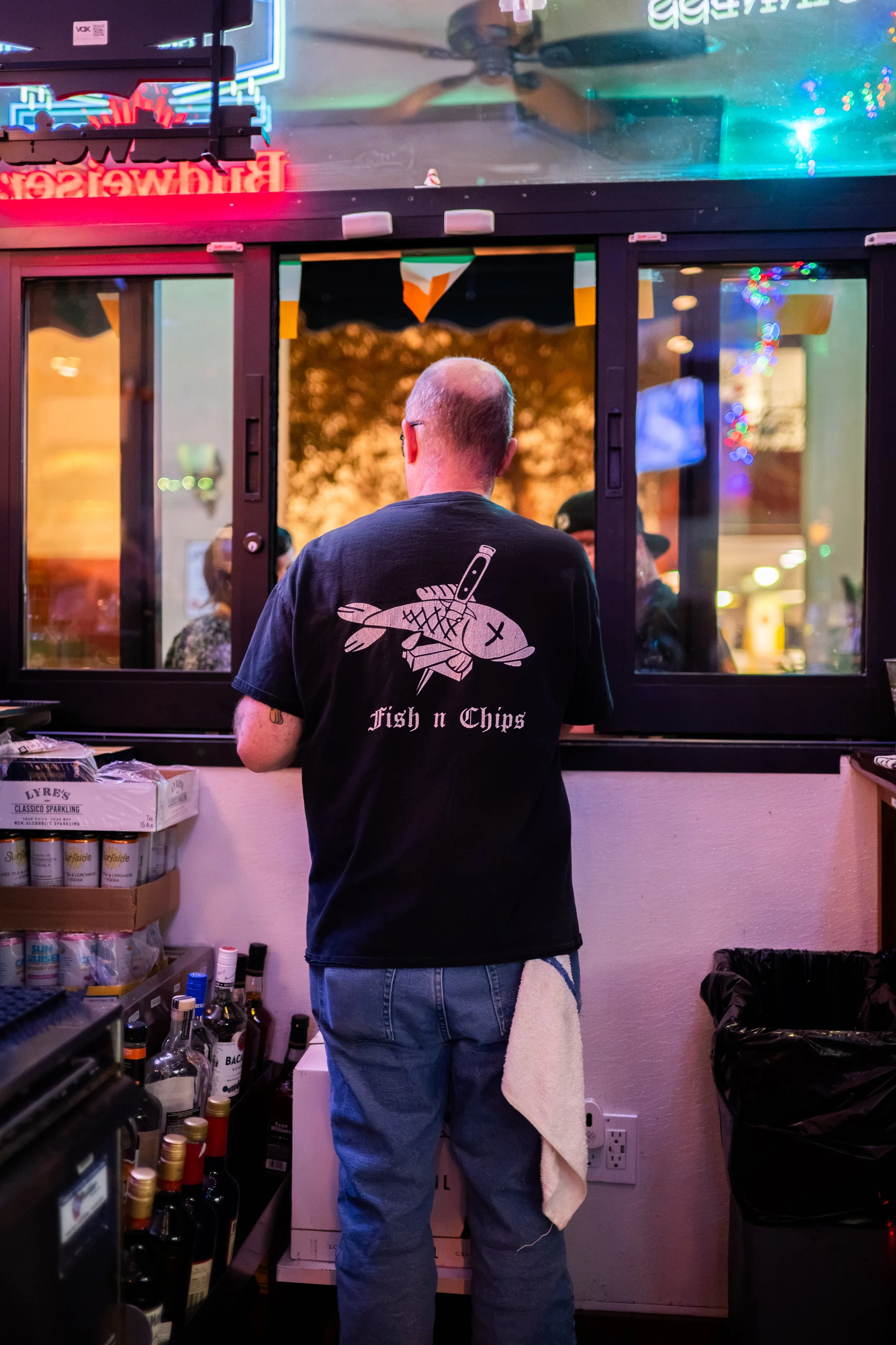 A man wearing a black t-shirt with a fish and knife graphic and the words 'Fish n Chips' on the back, standing in front of a window at a counter with bottles of alcohol. He has a towel hanging from his pocket and is facing away from the camera. The b