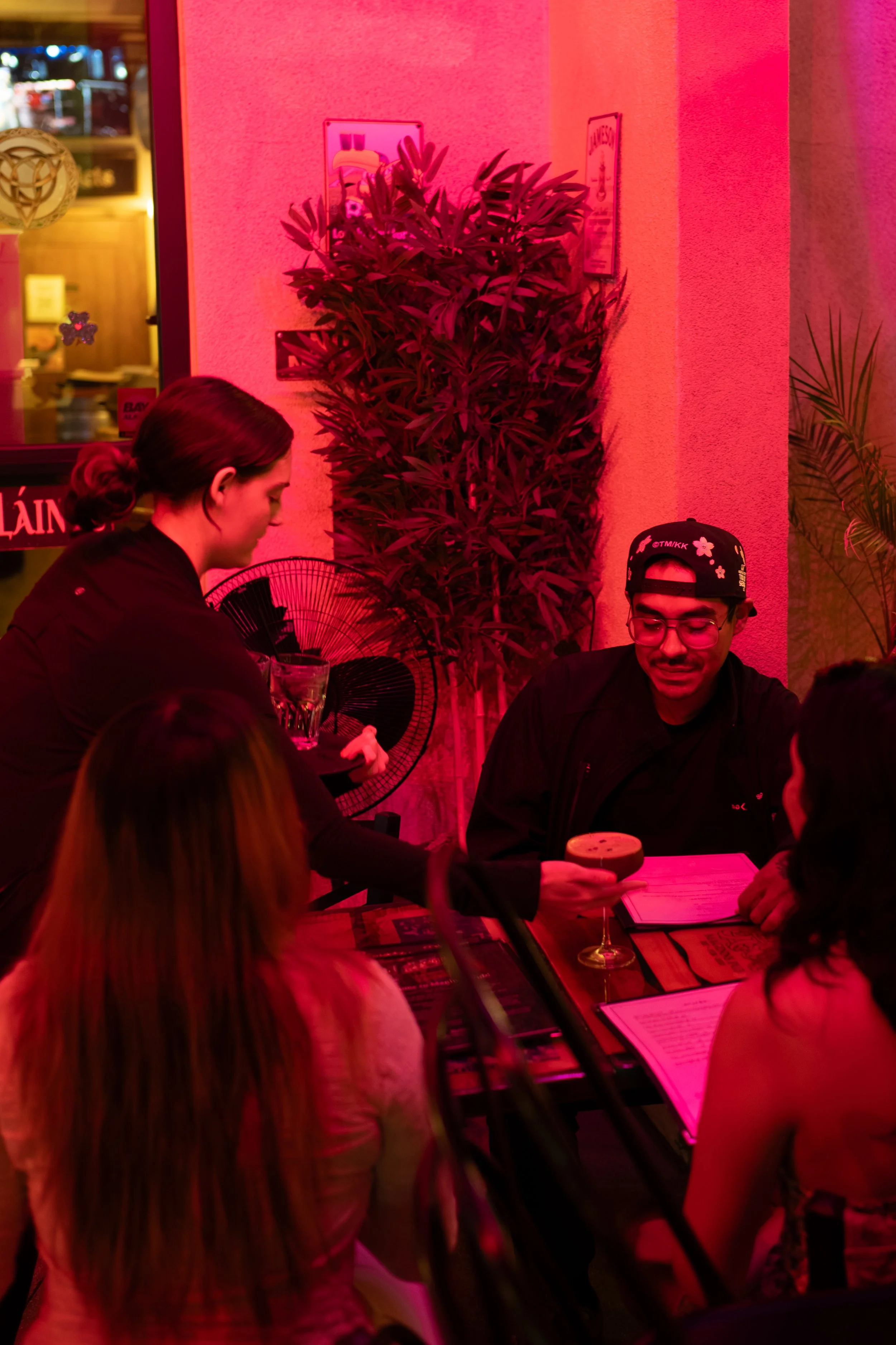 A group of people sitting at a table in a restaurant or bar with pink lighting, with two women standing and talking to the seated men, one of whom is holding a drink and a menu.
