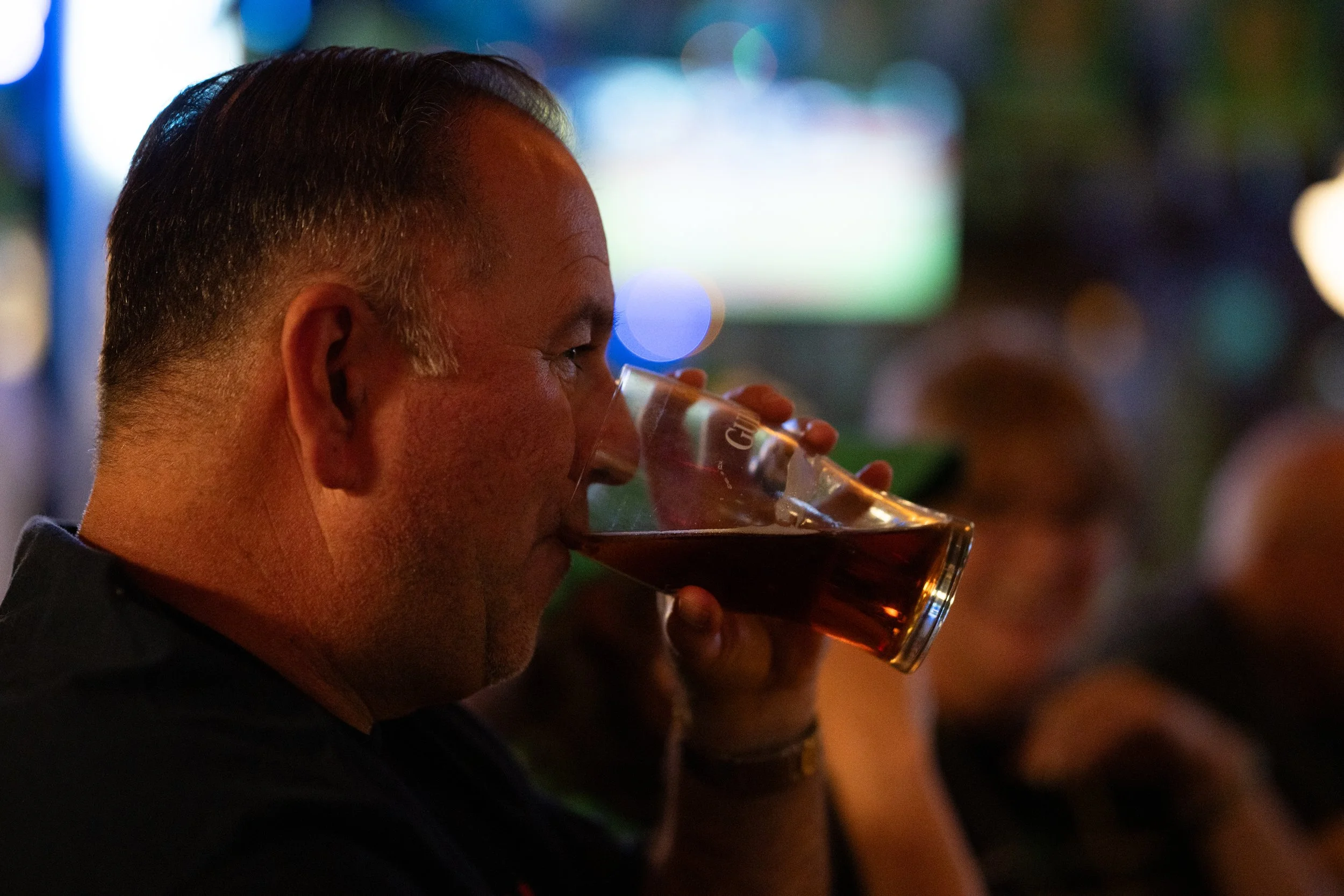 A man with short gray hair drinks a dark beer from a glass in a dimly lit bar or pub.