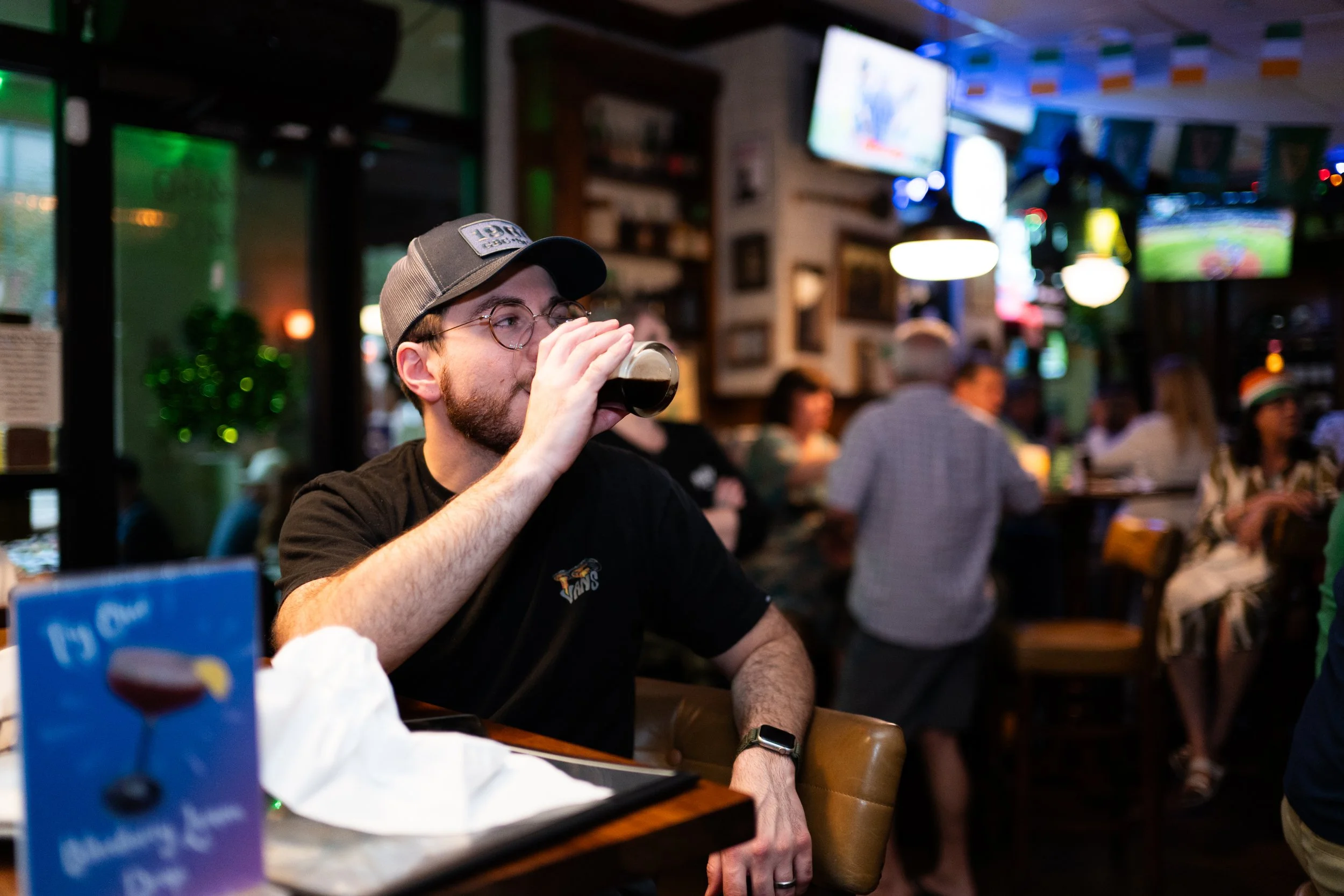 A man drinking a dark beer in a lively bar with multiple television screens and other patrons in the background