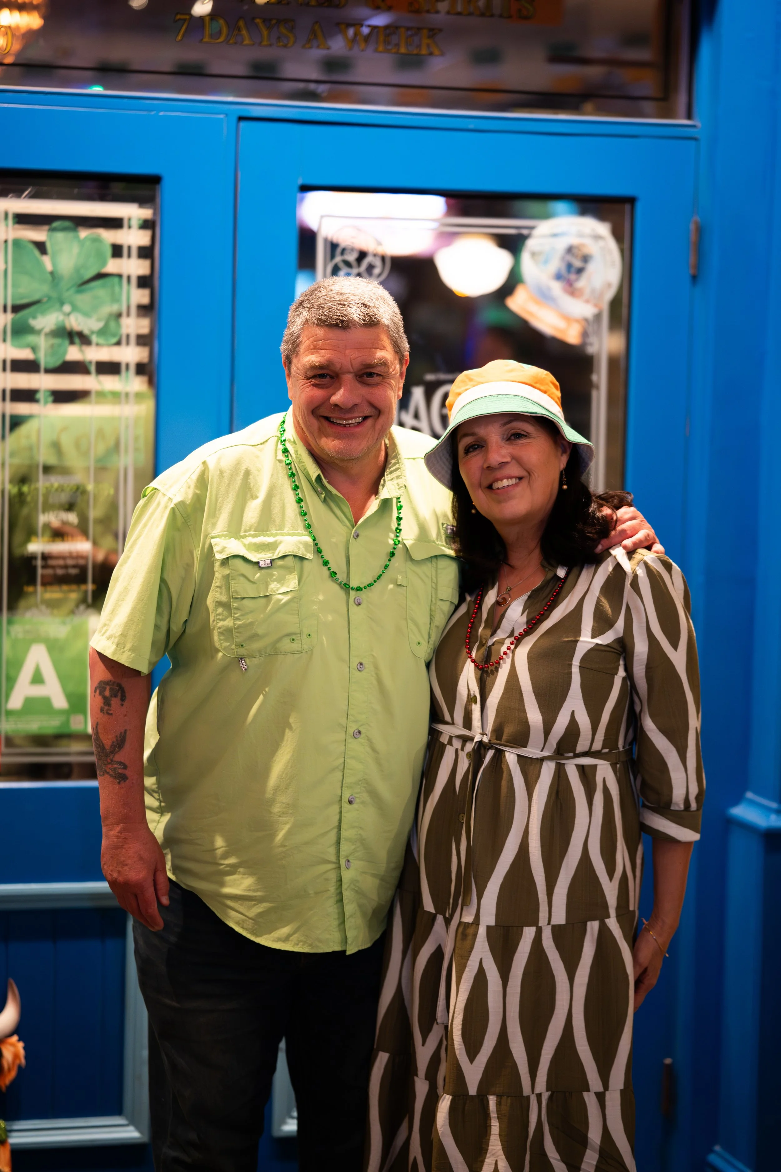 A smiling man and woman standing close together in front of a blue door, with the man wearing a light green shirt and the woman dressed in a patterned brown and white dress, both wearing colorful beads and the woman also wearing a hat.