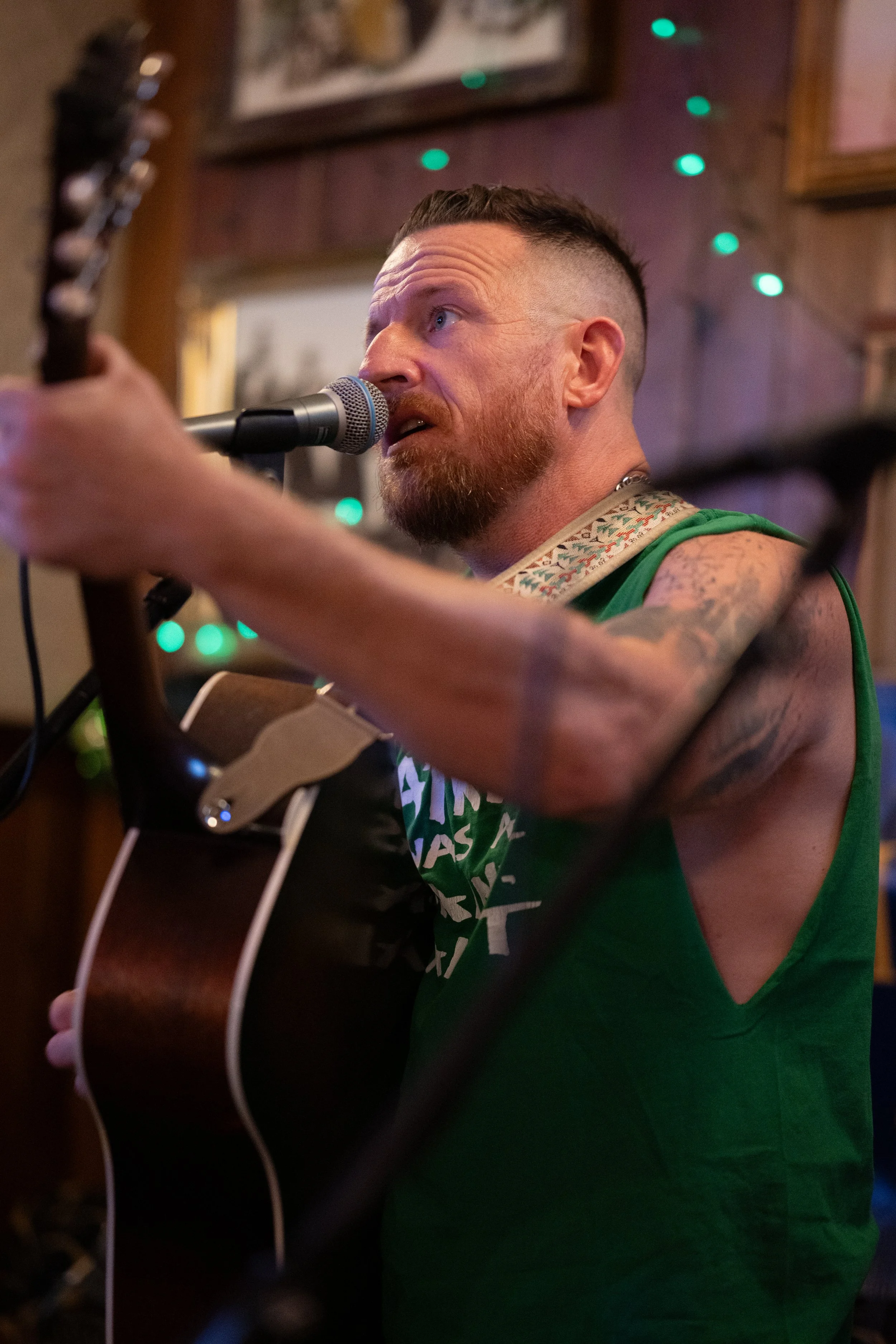 A man with a beard and tattoos playing an acoustic guitar and singing into a microphone in a cozy, dimly lit room decorated with green string lights.