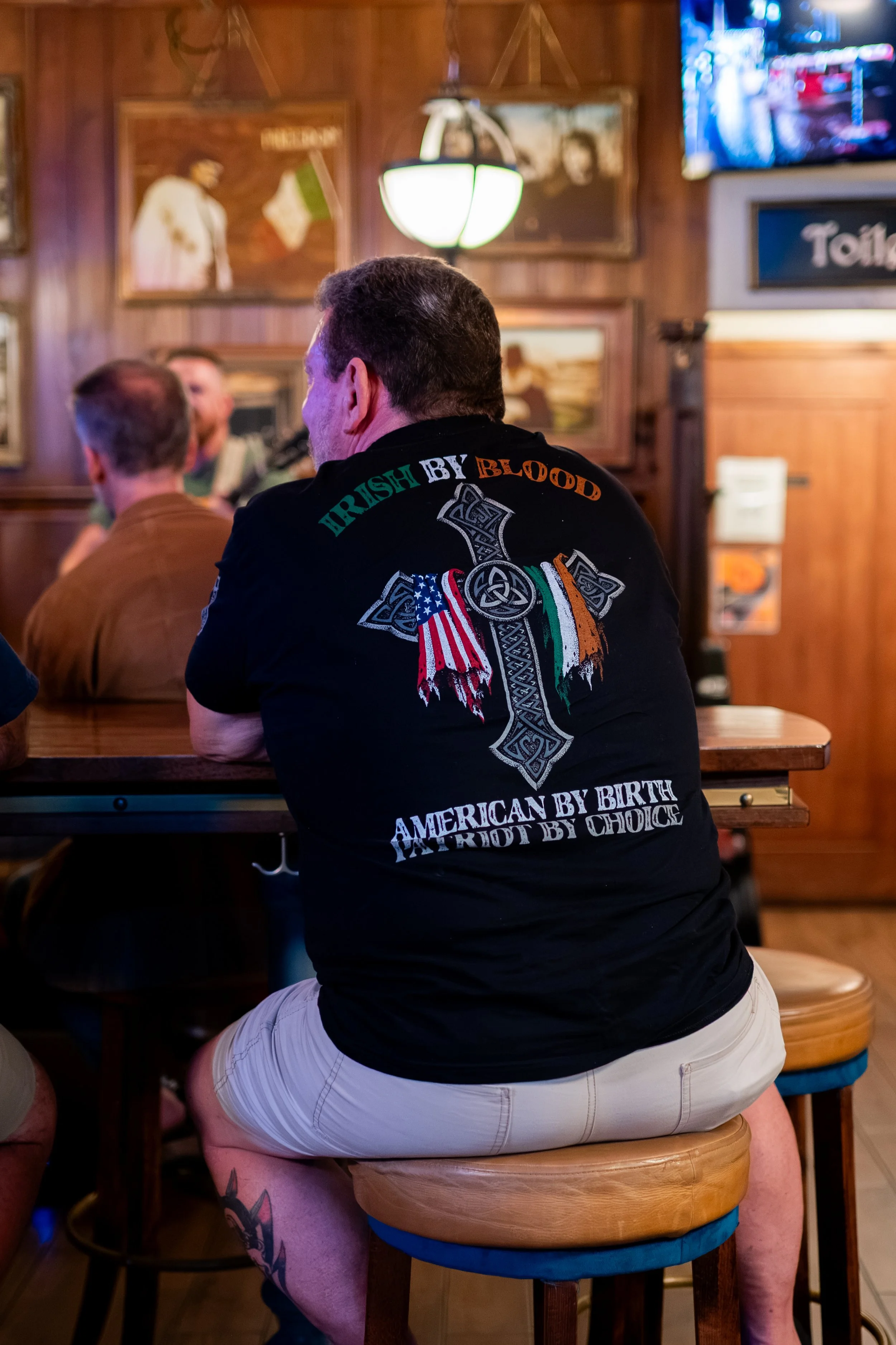A man with tattoos on his leg wearing a black T-shirt with a Celtic cross and patriotic words, sitting on a barstool in a bar with wooden walls, framed pictures, and a television.