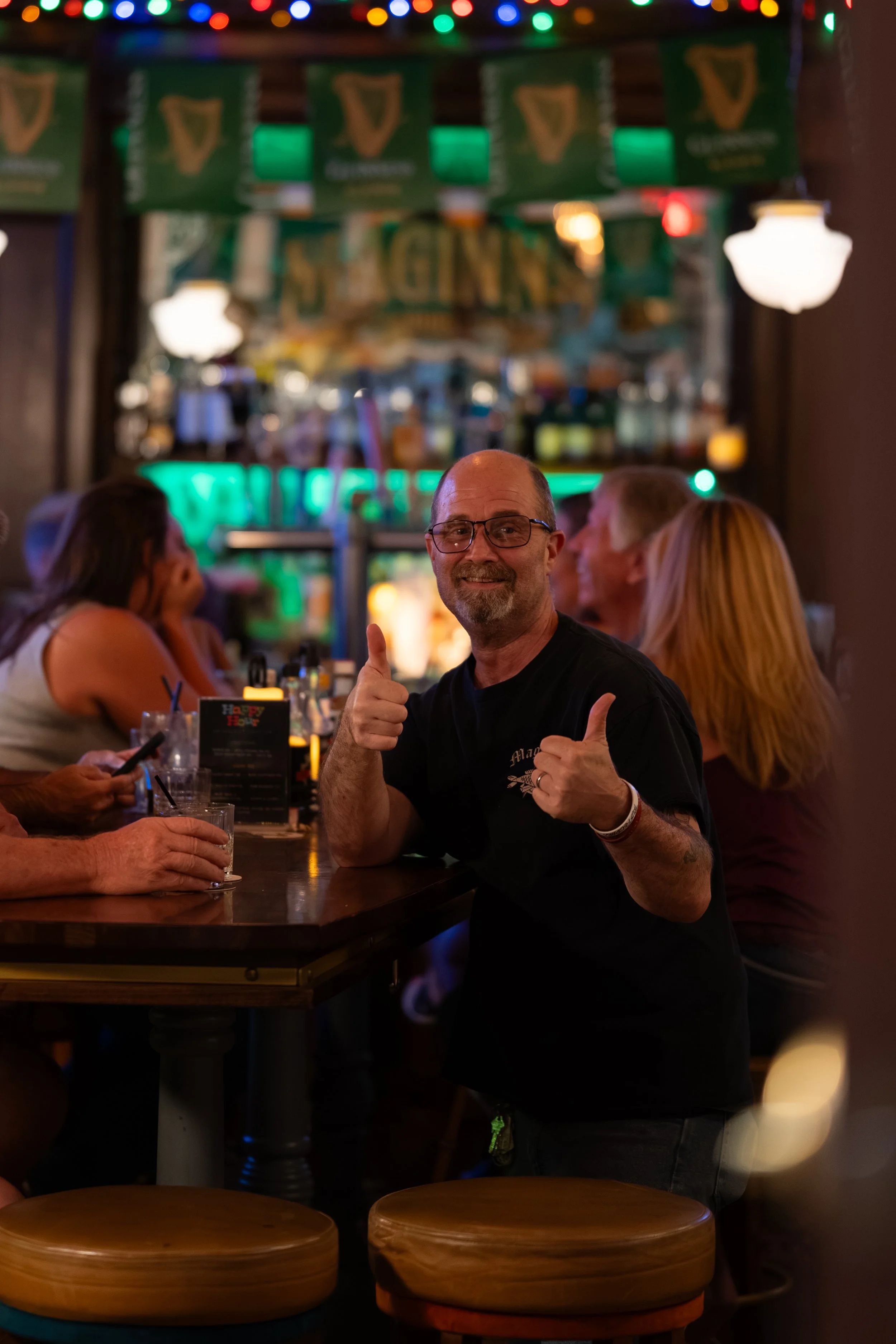 A man with glasses smiling and giving a thumbs-up in a dimly lit restaurant or bar. He is surrounded by other people, some of whom are talking or looking at their phones. The background features colorful lights and a bar area.